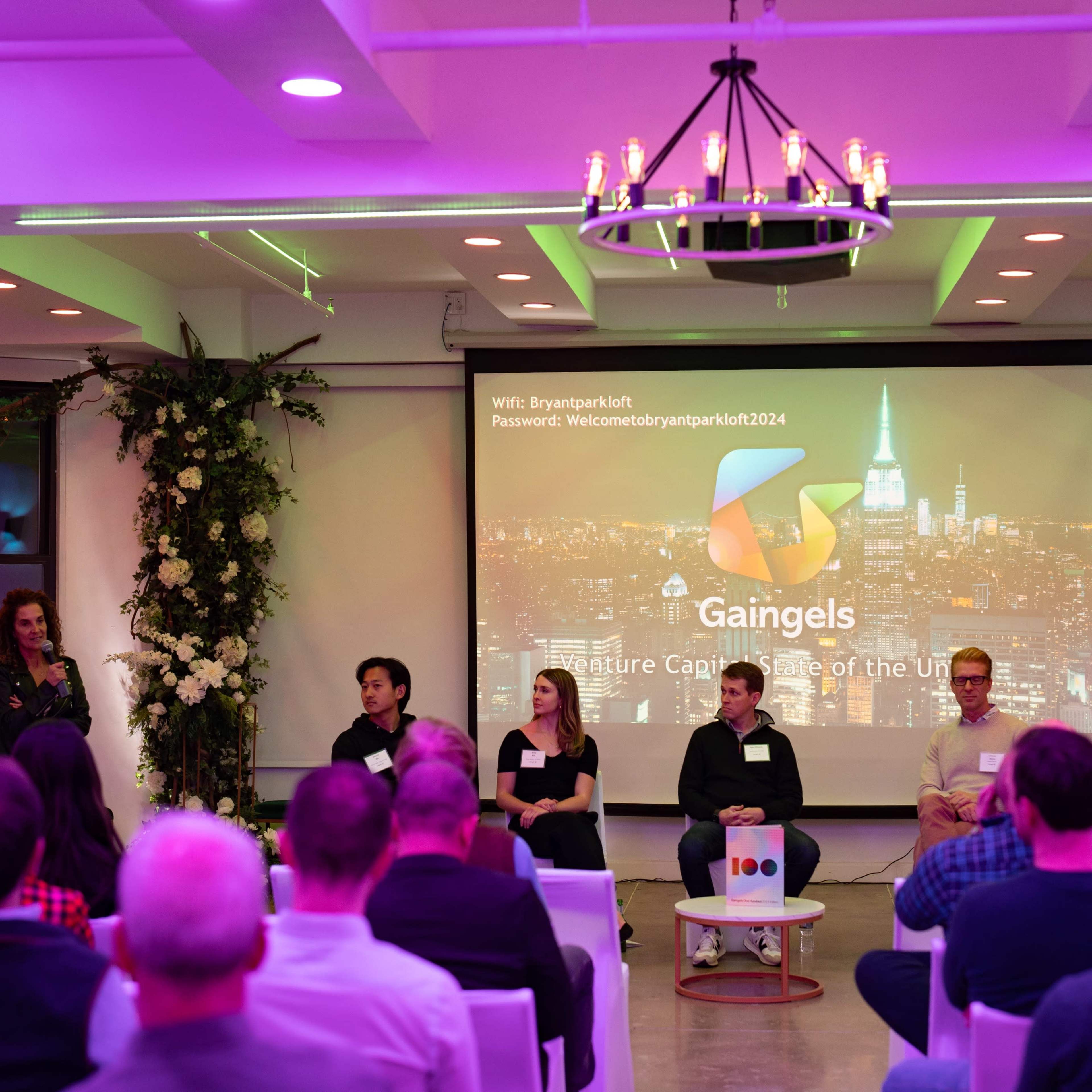 A panel discussion takes place in a well-lit venue with a large screen displaying the Gaingels logo and a cityscape backdrop.