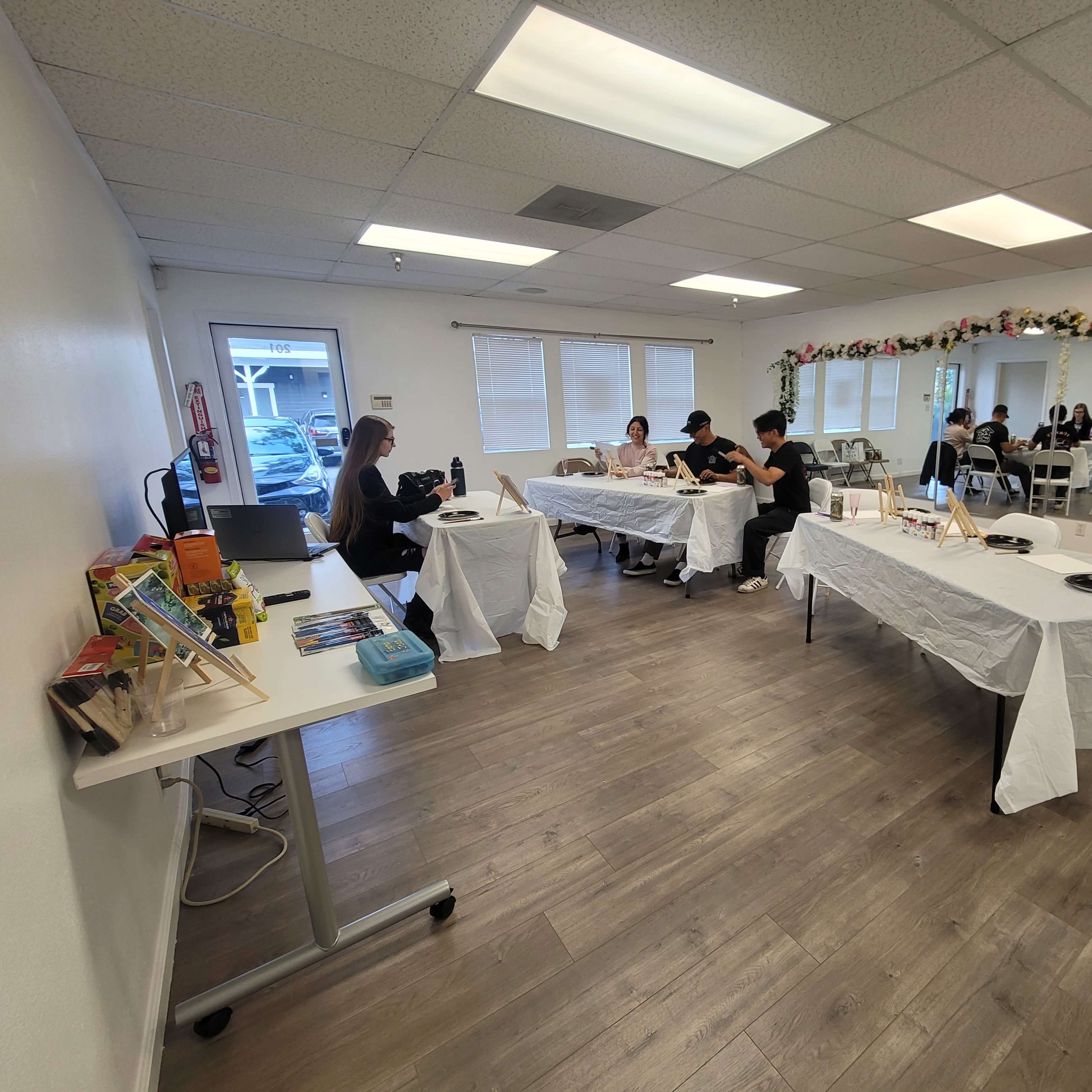 A spacious, well-lit room with several tables covered in white cloth, where people are engaged in activities, surrounded by decorative elements along the walls.