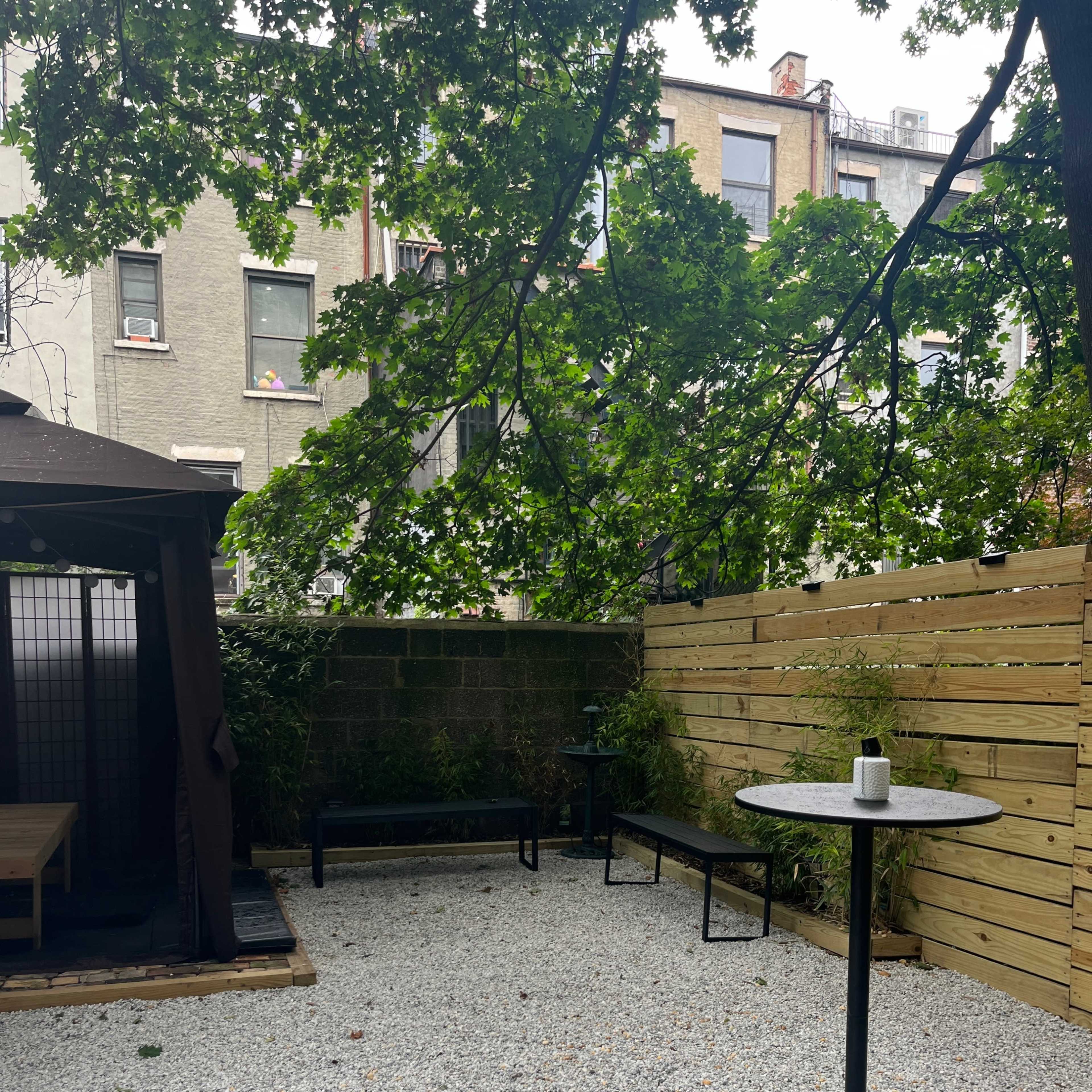 The scene features a gravel courtyard surrounded by wooden fencing and greenery, with a table and chairs under a large tree.