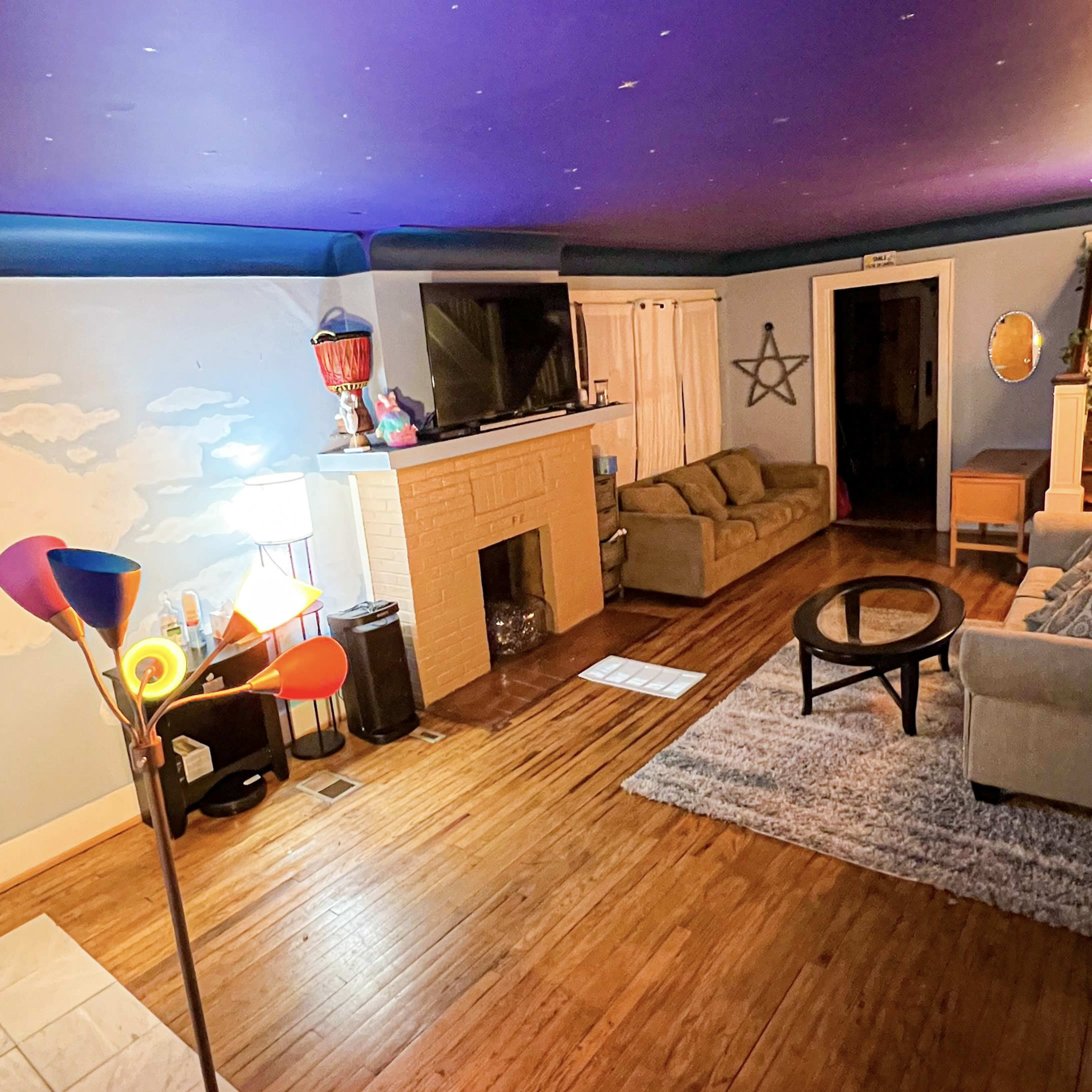 A living room with a purple ceiling, a light-colored wall featuring a cloud mural, a fireplace, two couches, a round coffee table, and various light sources.