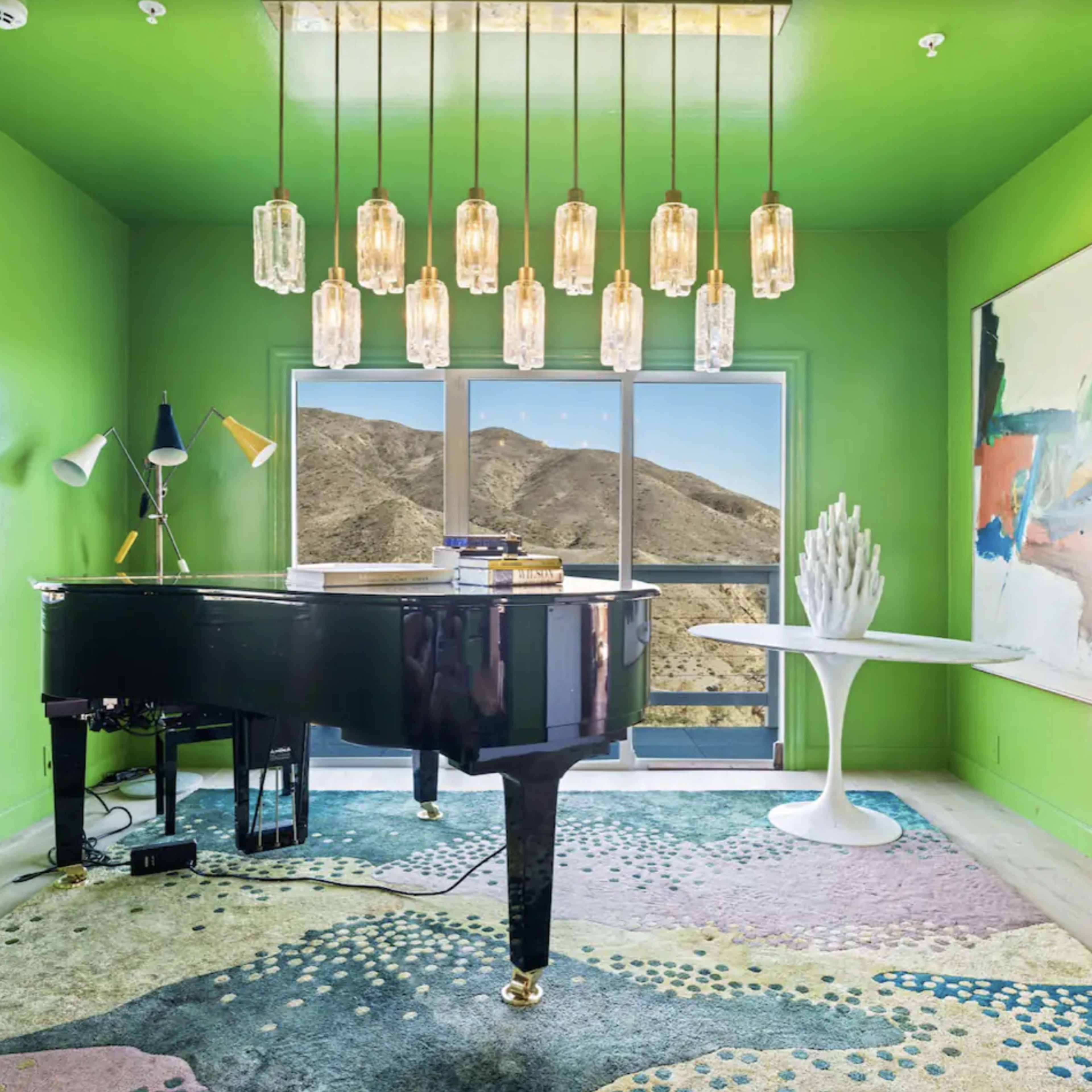 A grand piano is placed in a brightly colored green room with large windows showcasing a mountain view, illuminated by pendant lights and accompanied by modern artwork and a patterned rug.