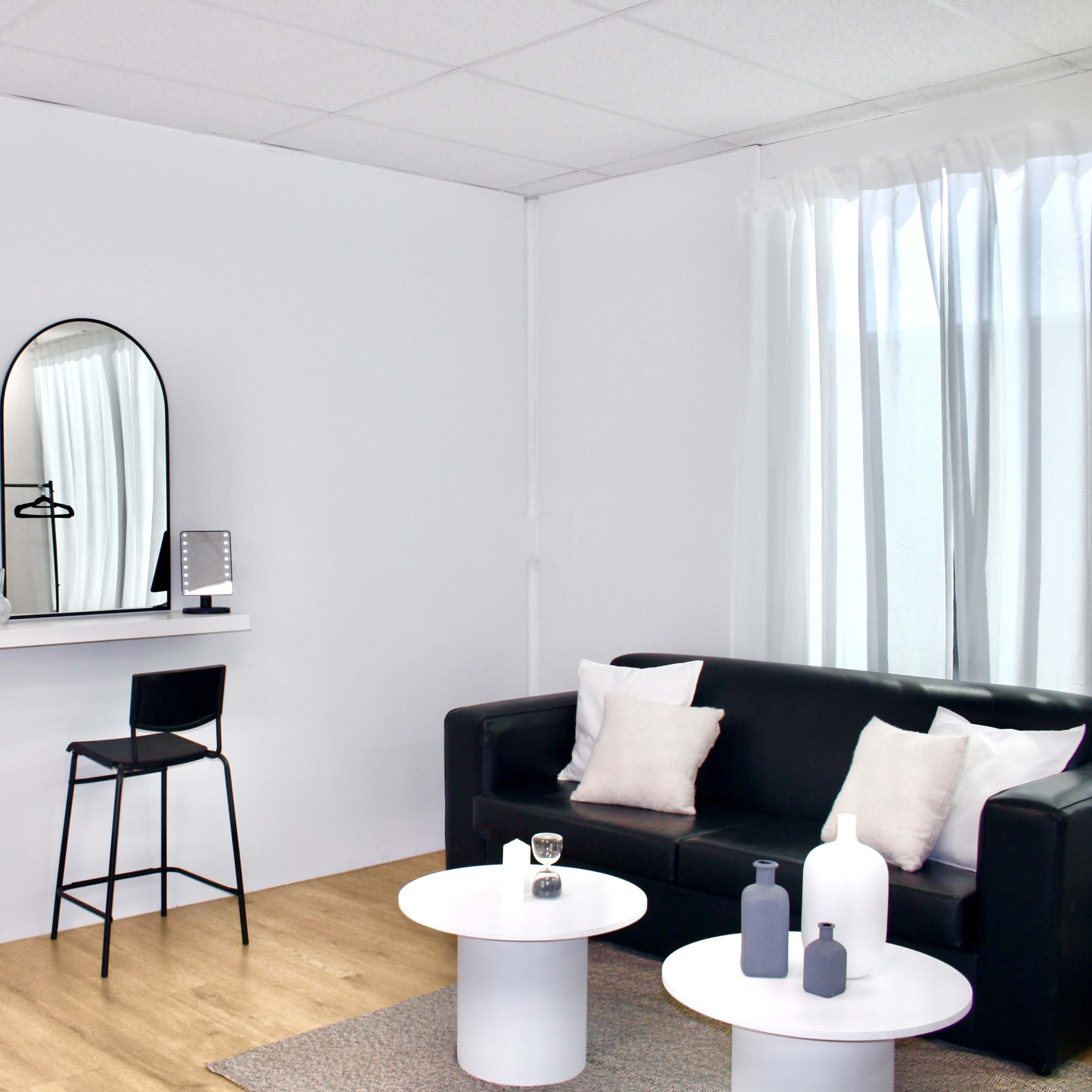 A modern living space featuring a black sofa, two black bar stools at a wall-mounted shelf with mirrors above, and two round white tables adorned with decorative items.