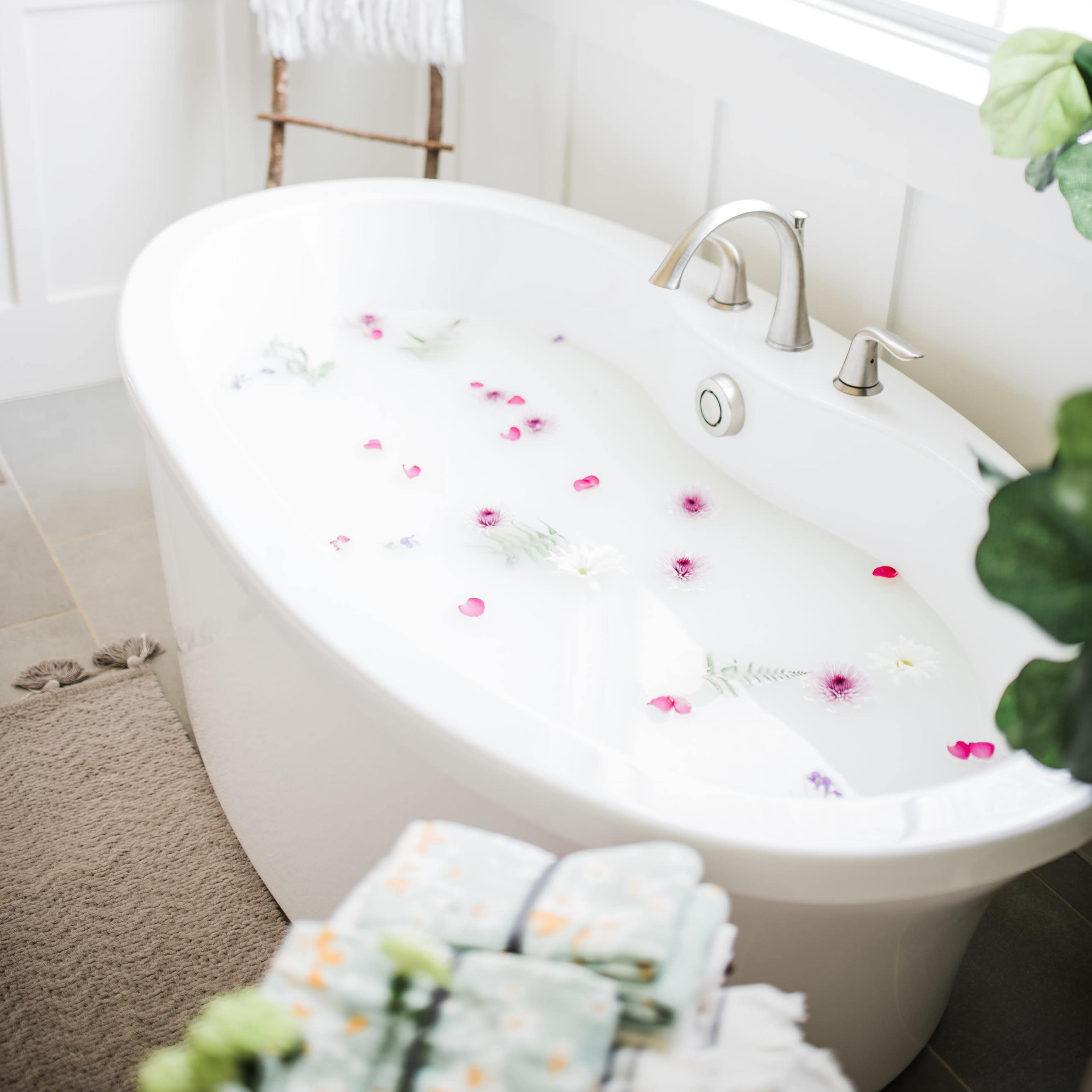 A modern freestanding bathtub filled with water and flower petals is positioned beside a window, with a ladder holding a towel nearby.