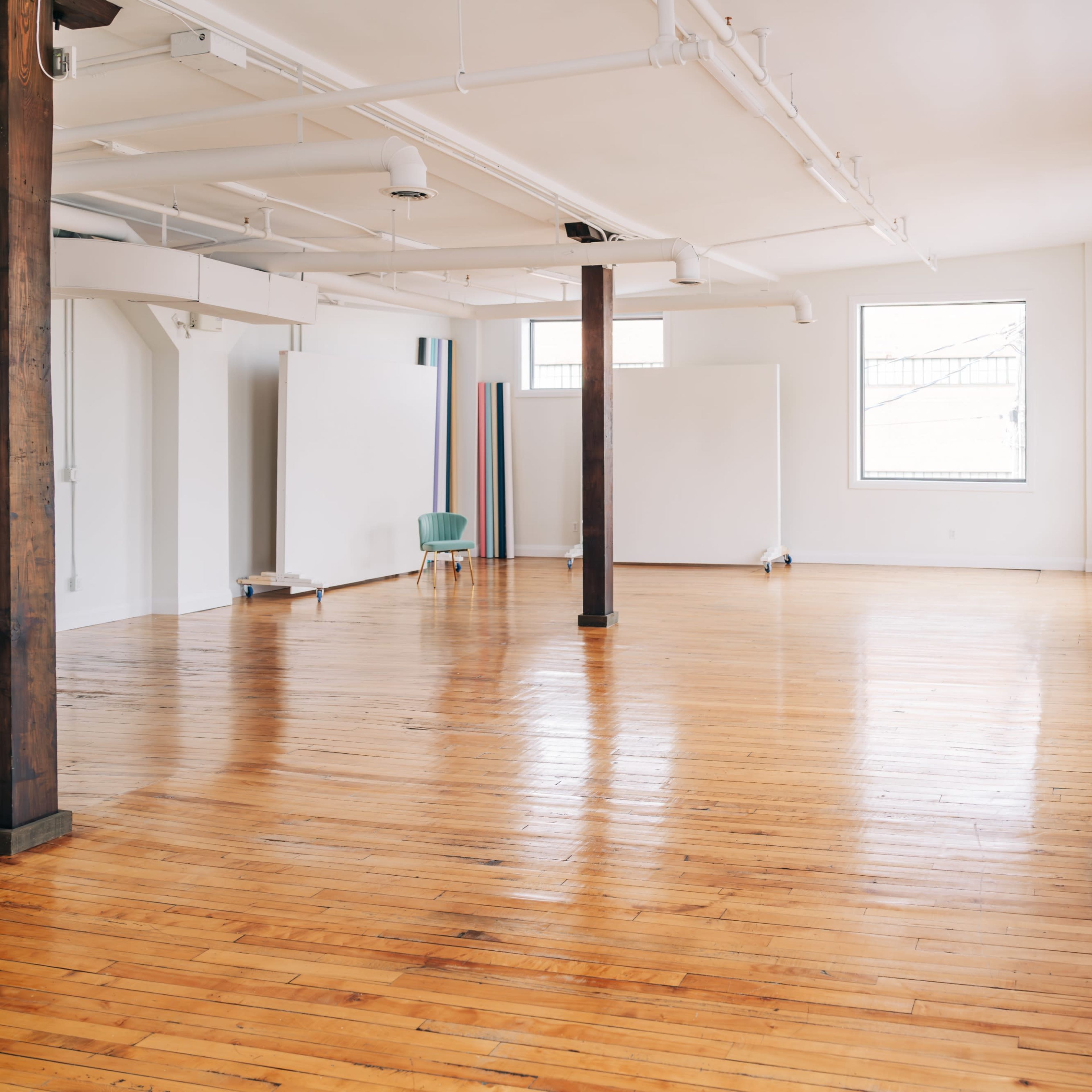 A spacious, empty room with hardwood floors, large windows, and a few pieces of minimalist furniture against the walls.