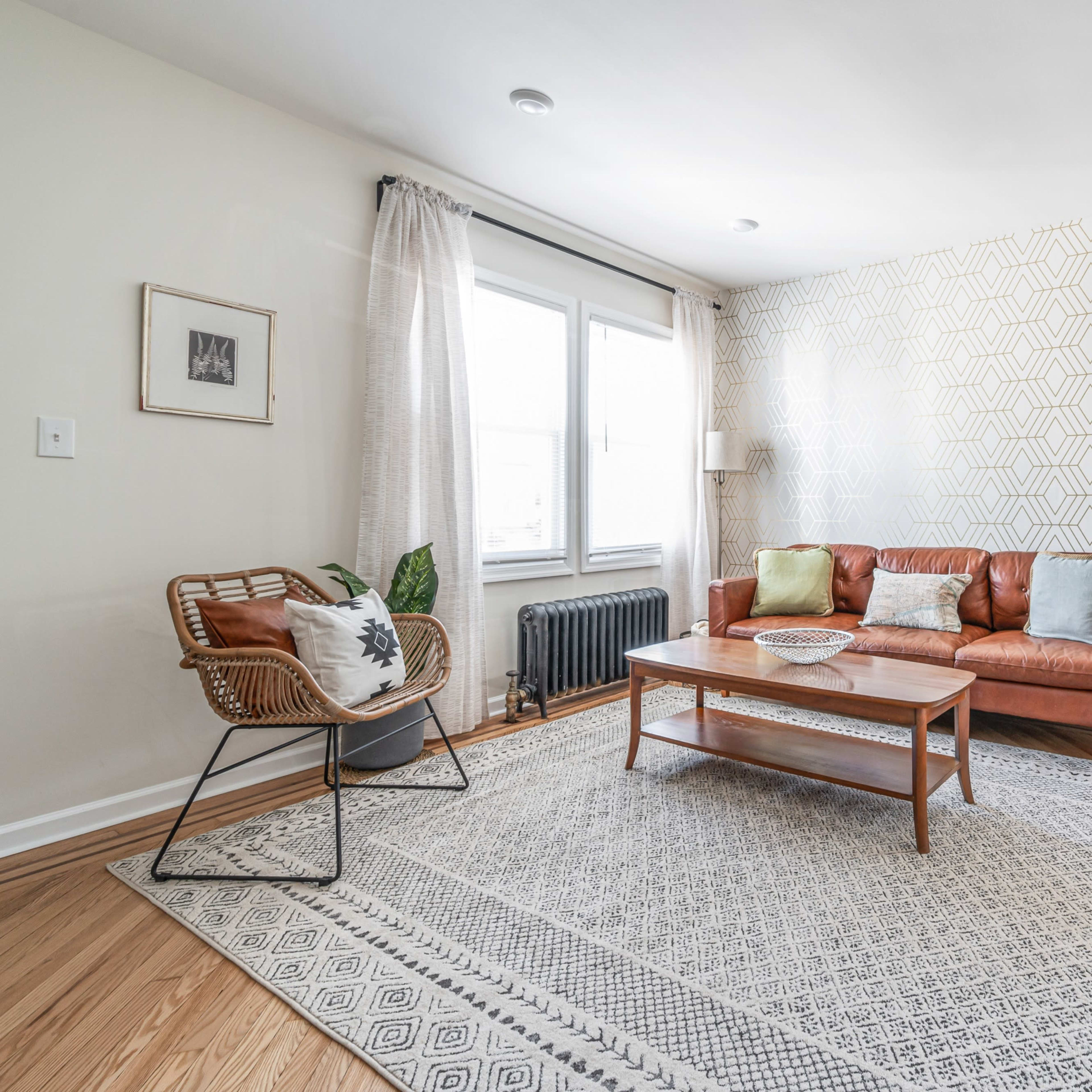 A bright living room featuring a leather sofa, a patterned rug, a wooden coffee table, and a large window with sheer curtains.