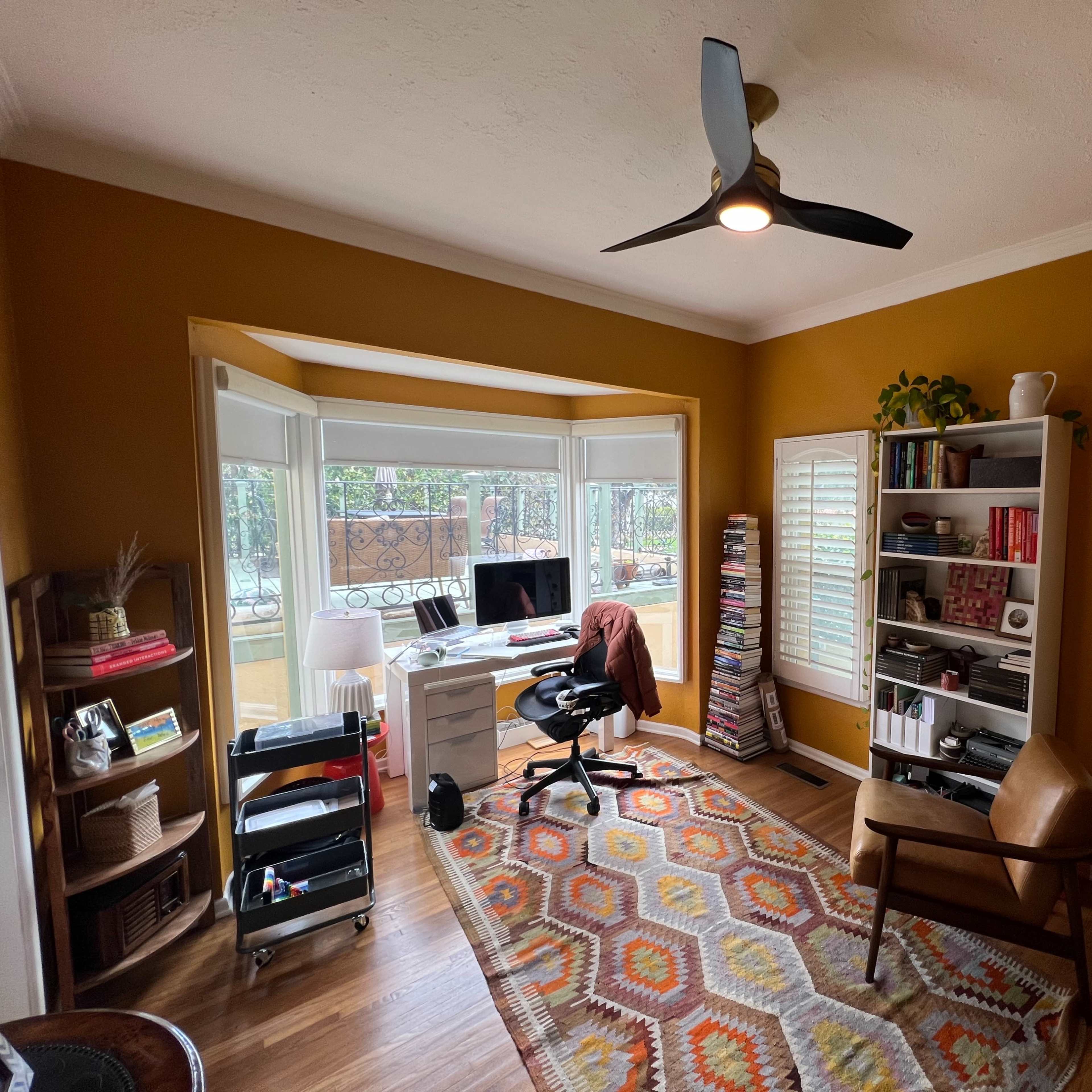 A home office with a desk, a black office chair, a brown armchair, shelves filled with books, and large windows providing natural light.