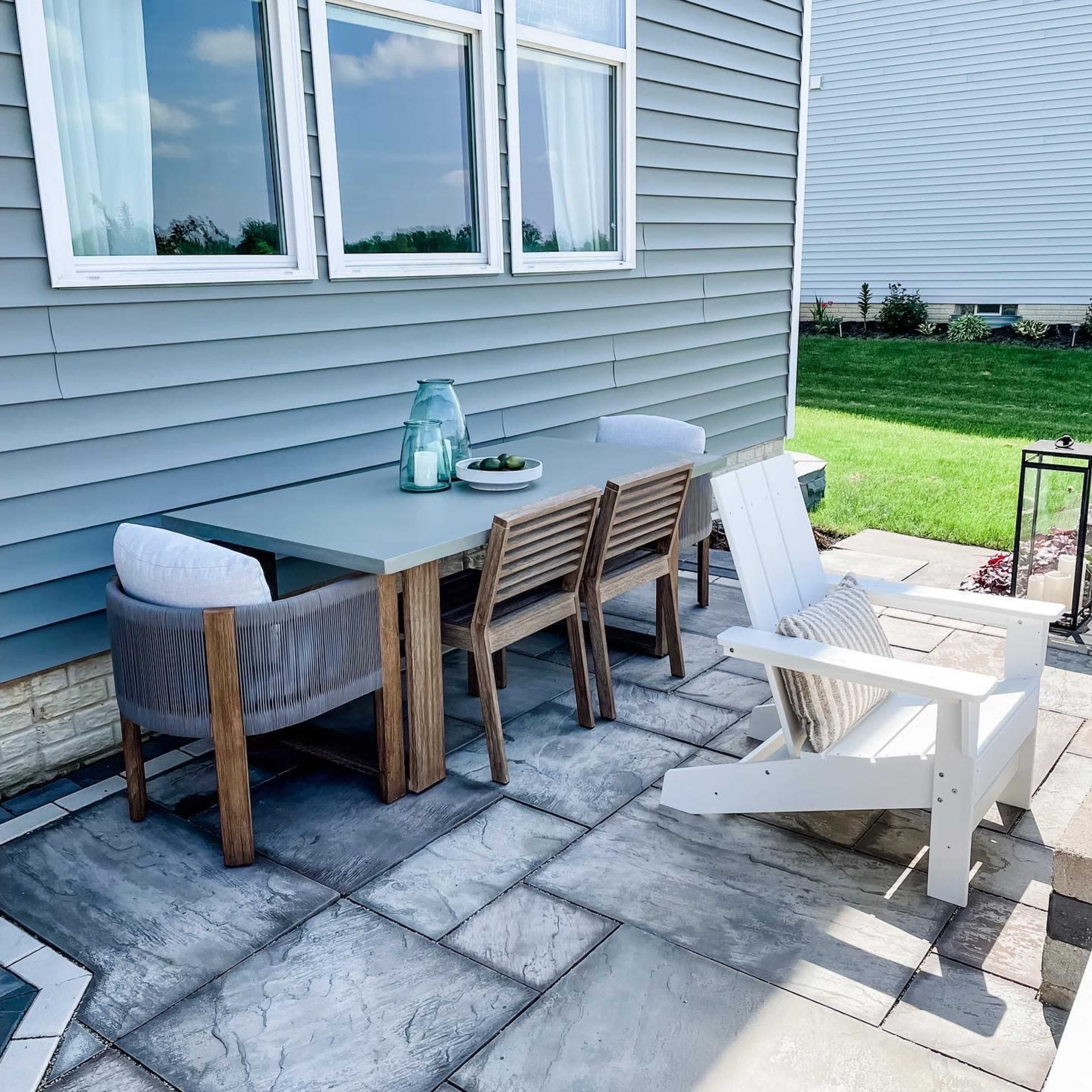 A patio area features a rectangular table with four wooden chairs and an Adirondack chair, situated on stone paving beside a house with gray siding.