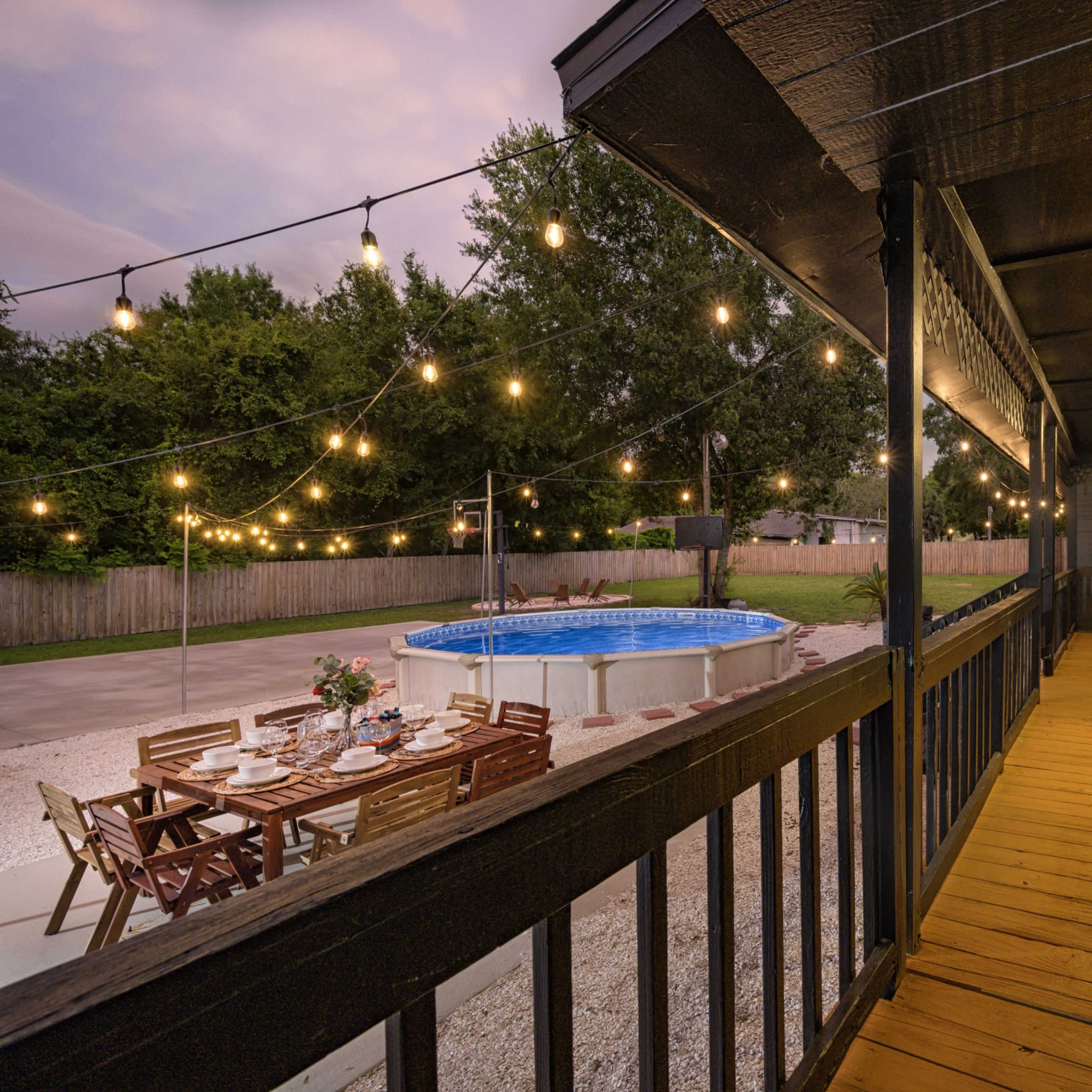 A wooden deck overlooks a backyard with a swimming pool, dining table set with chairs, and string lights overhead.