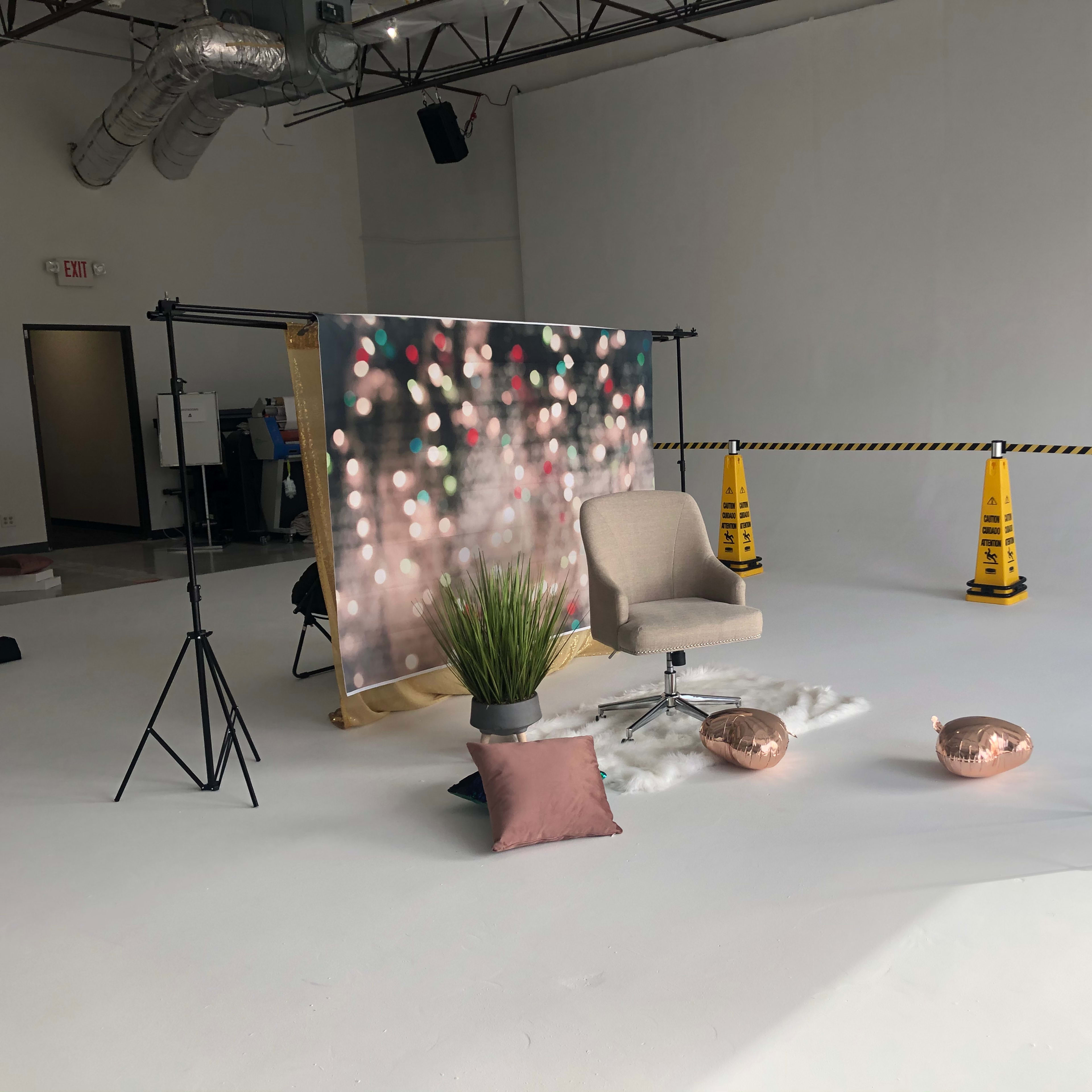 A photography setup with a backdrop of blurred lights, two chairs, a small plant, decorative pillows, and caution cones marking off the area.