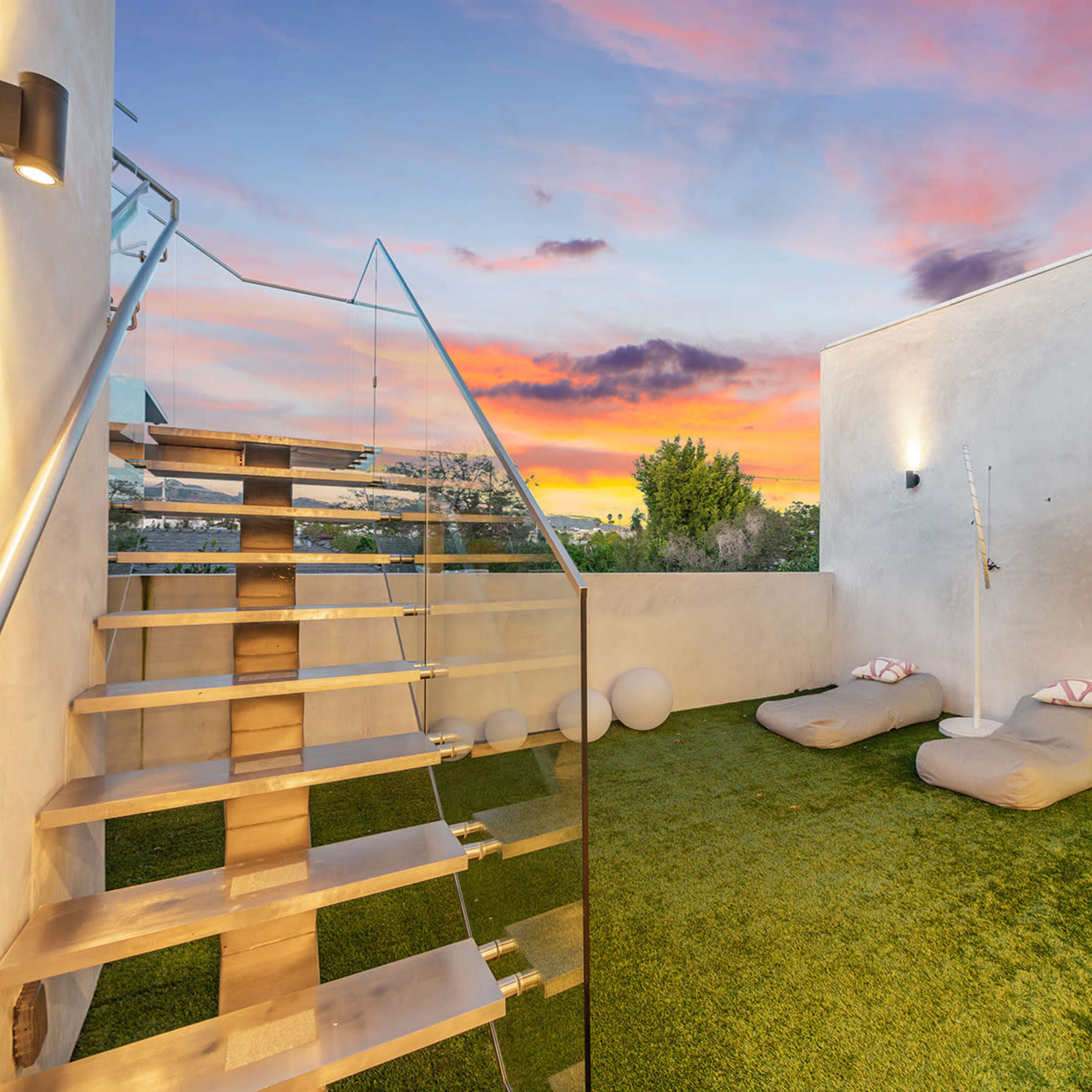 A modern outdoor space features a staircase leading to a grassy area with two lounge cushions and a colorful sunset in the background.