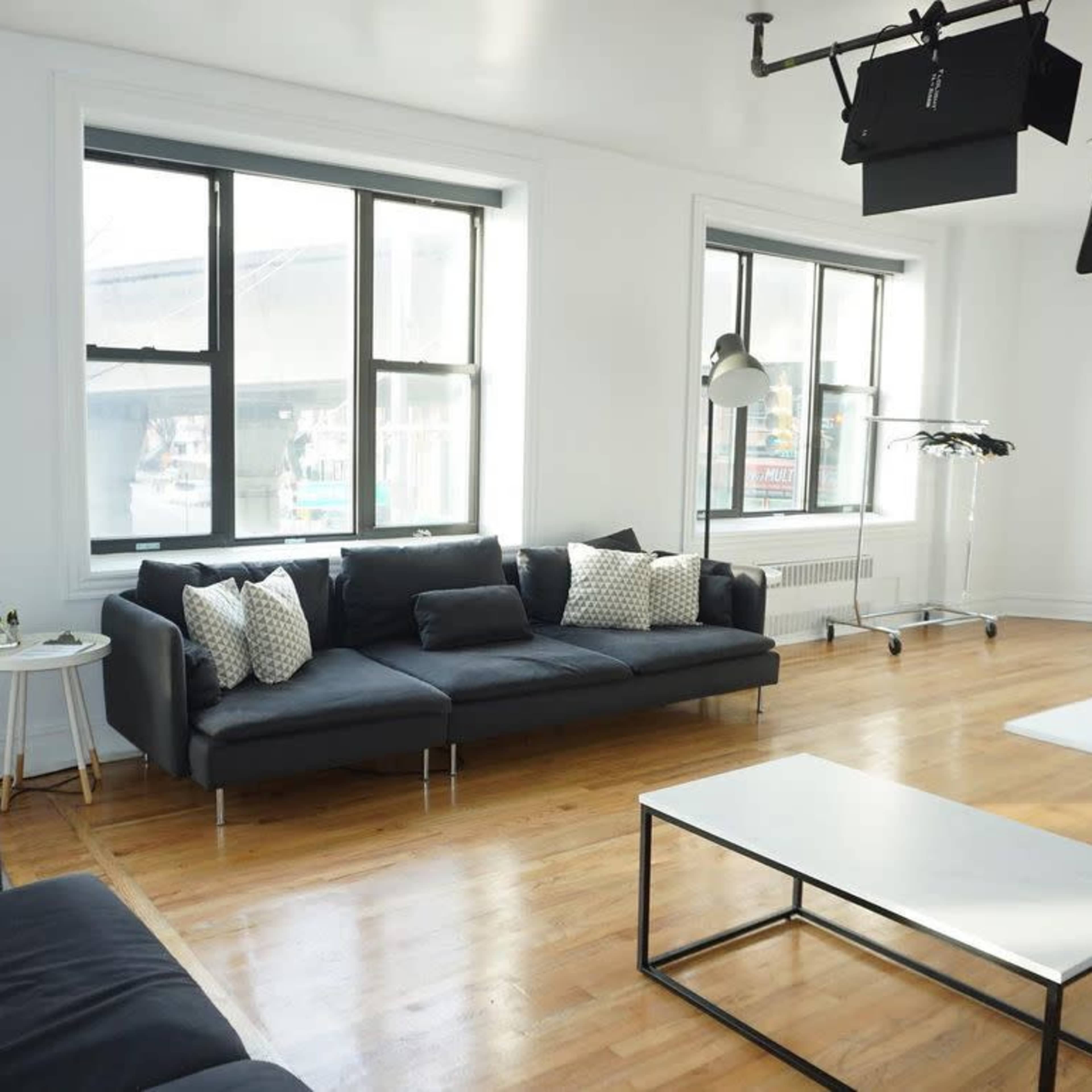 A bright, spacious room with large windows, a dark gray sectional sofa, a coffee table, and professional lighting equipment.
