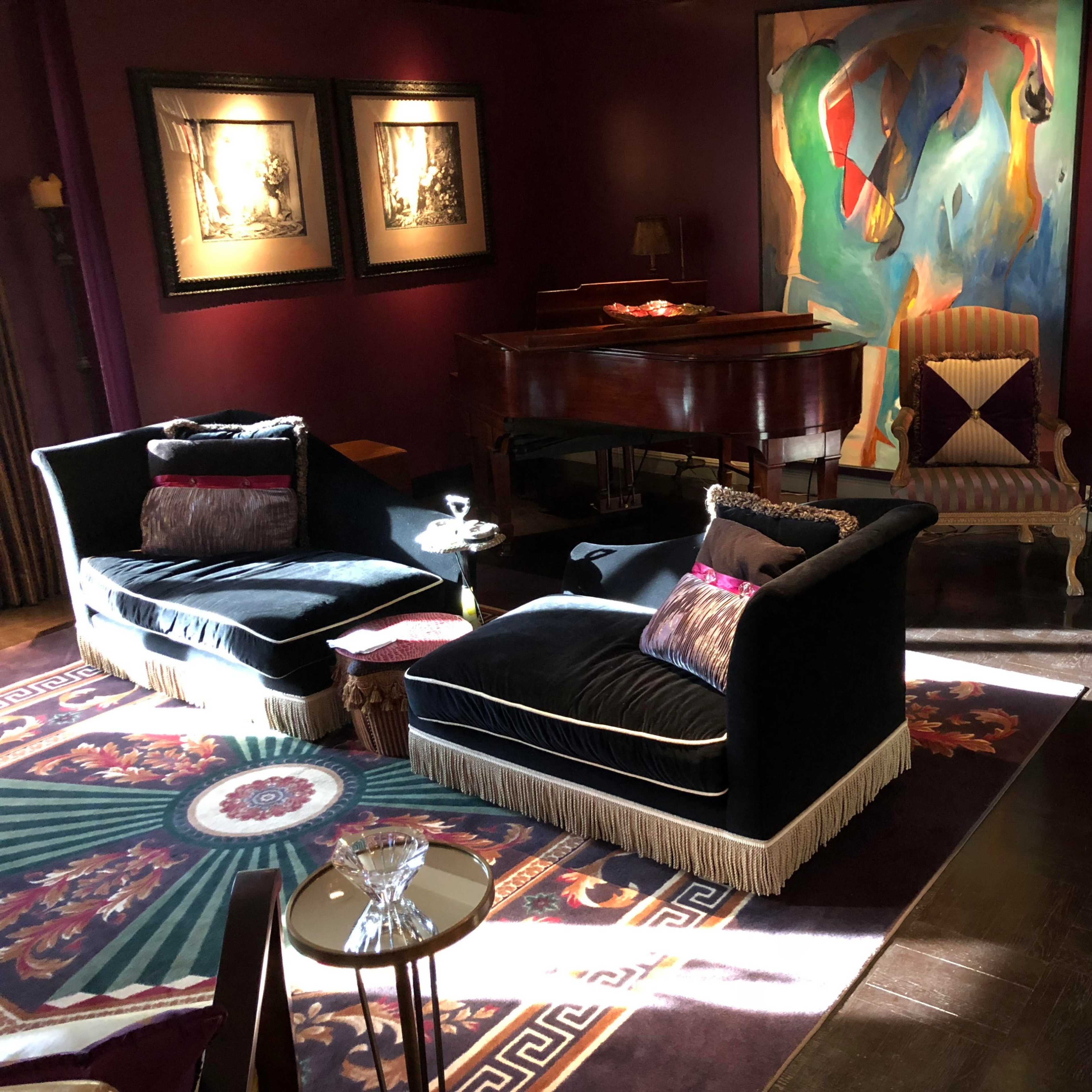 A cozy living room with two dark seating chairs, a grand piano, and colorful artwork on the walls, illuminated by natural light.
