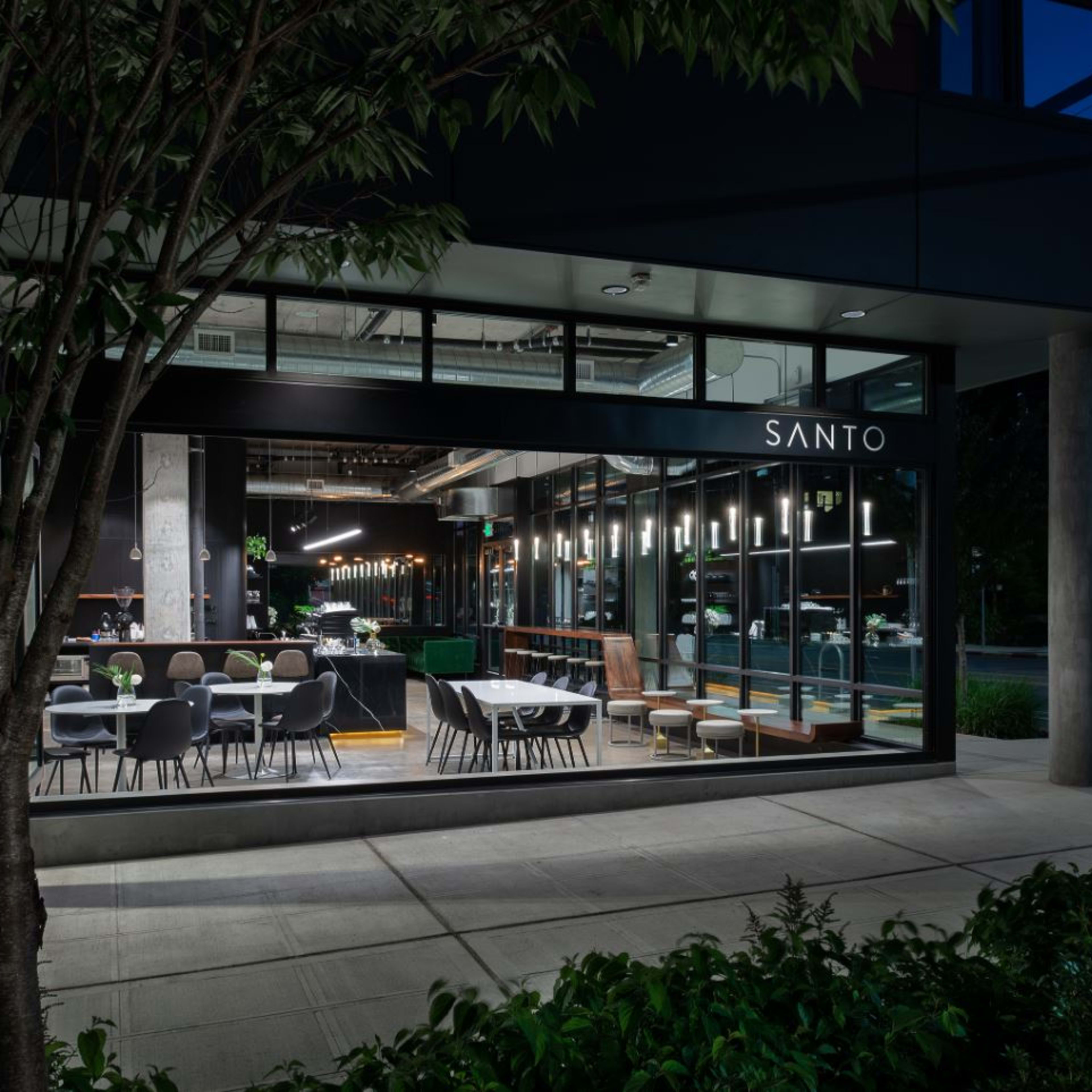 The exterior of a modern restaurant named "SANTO," with large windows displaying tables and seating arrangements, set against a nighttime backdrop.