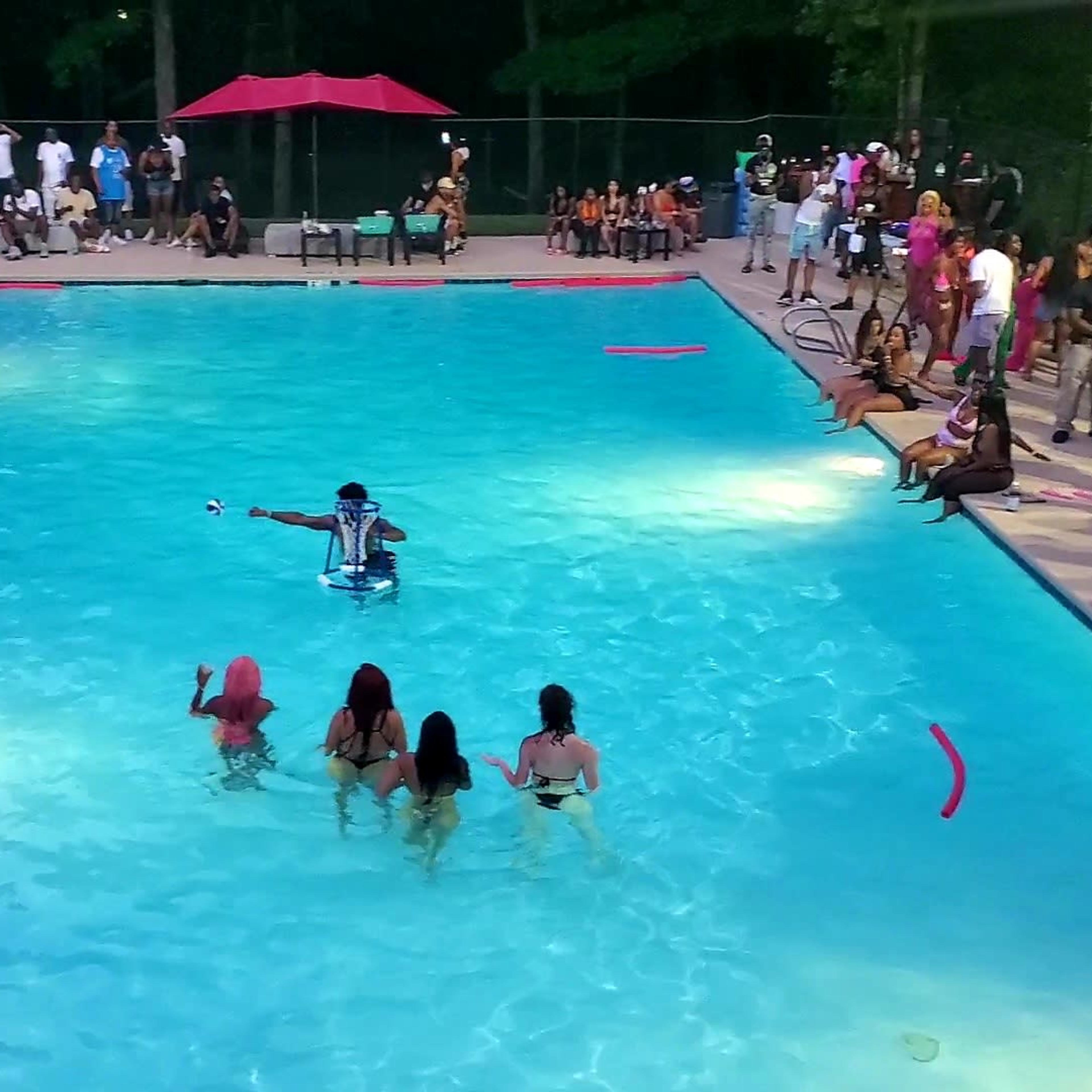 A lively outdoor pool scene features groups of people socializing and enjoying the water, with red and blue umbrellas providing shade.