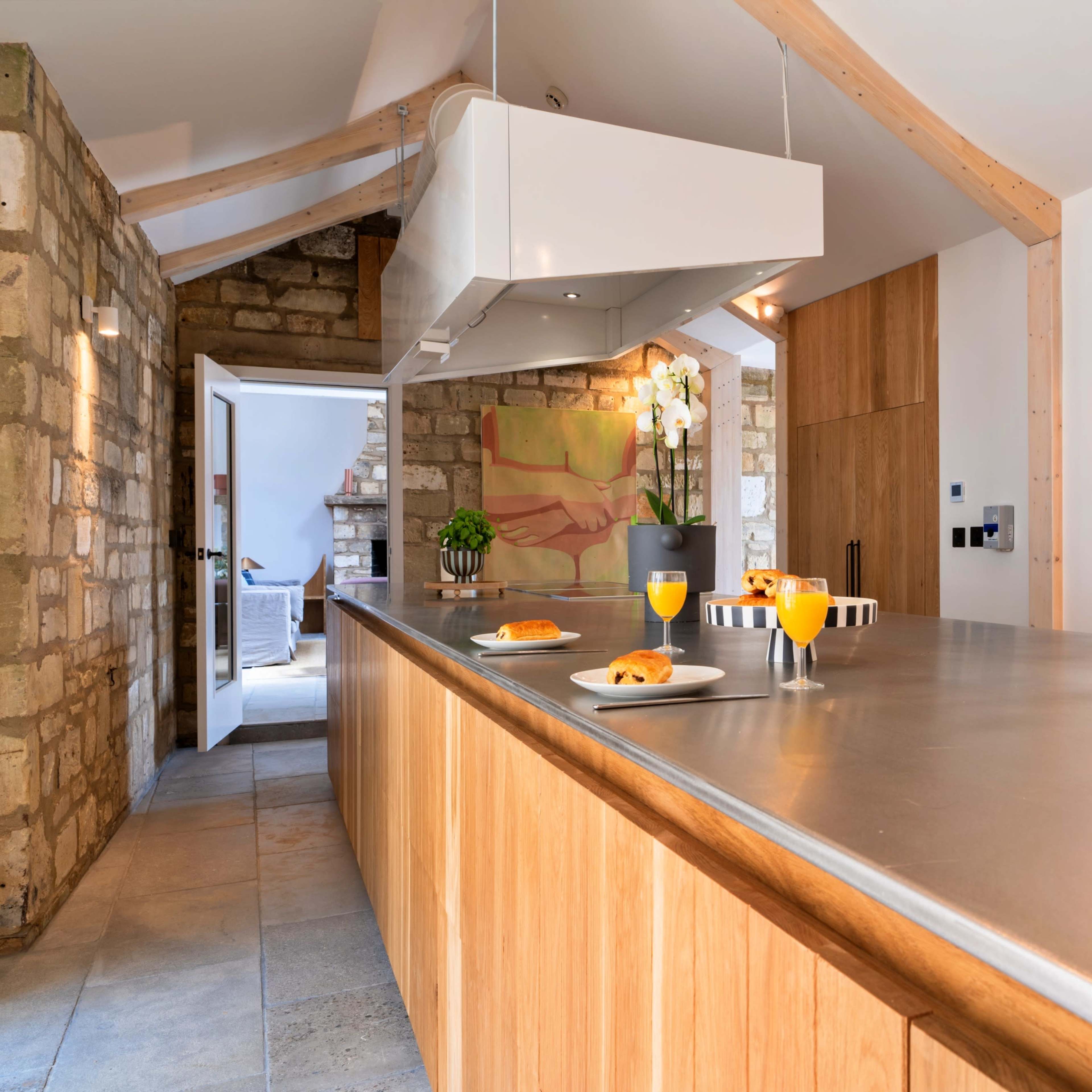 A spacious kitchen features a central wooden island with a metallic countertop, equipped with modern appliances and breakfast items, surrounded by stone and wooden accents.