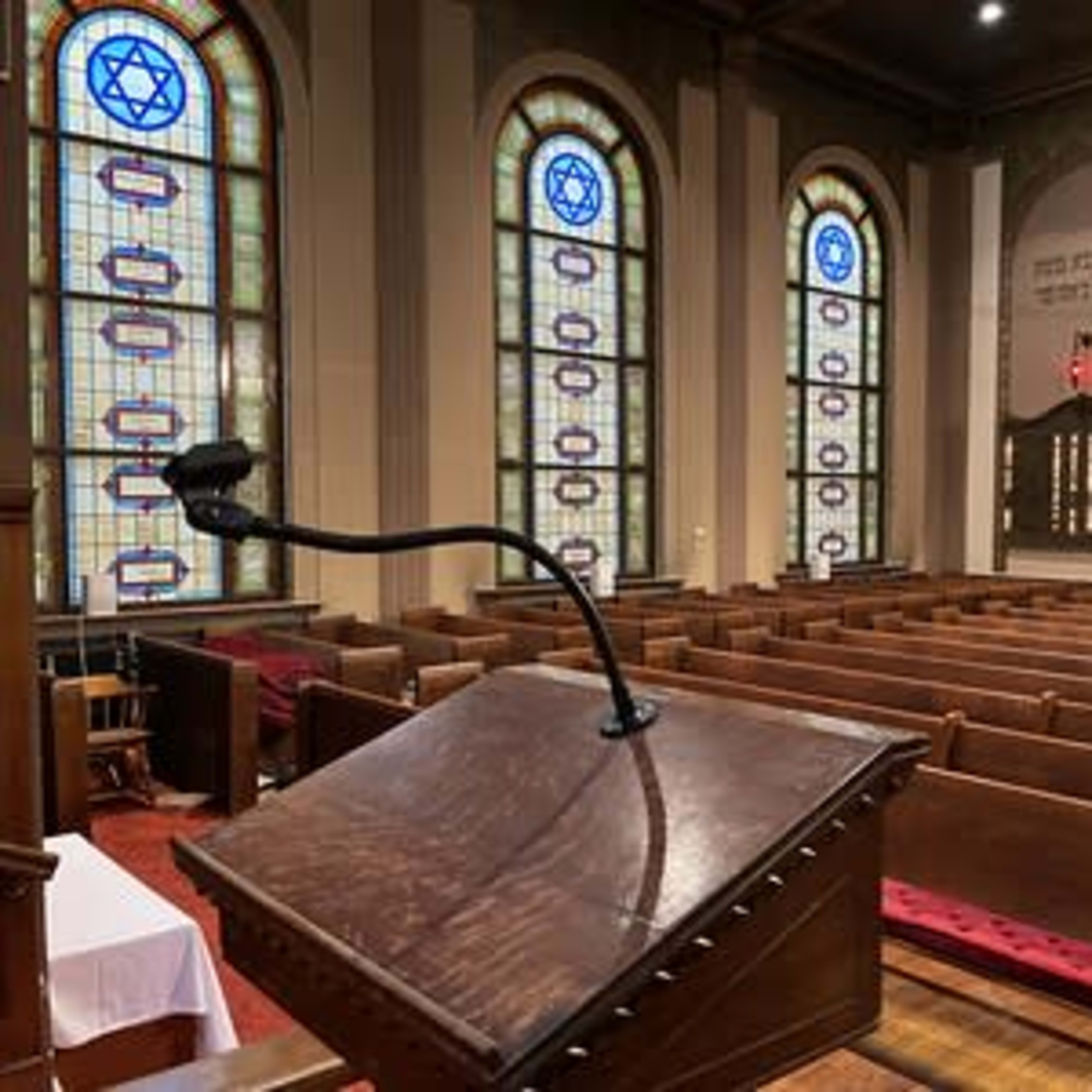 The interior of a synagogue with a wooden podium in the foreground, surrounded by rows of wooden benches and stained glass windows featuring Star of David symbols.