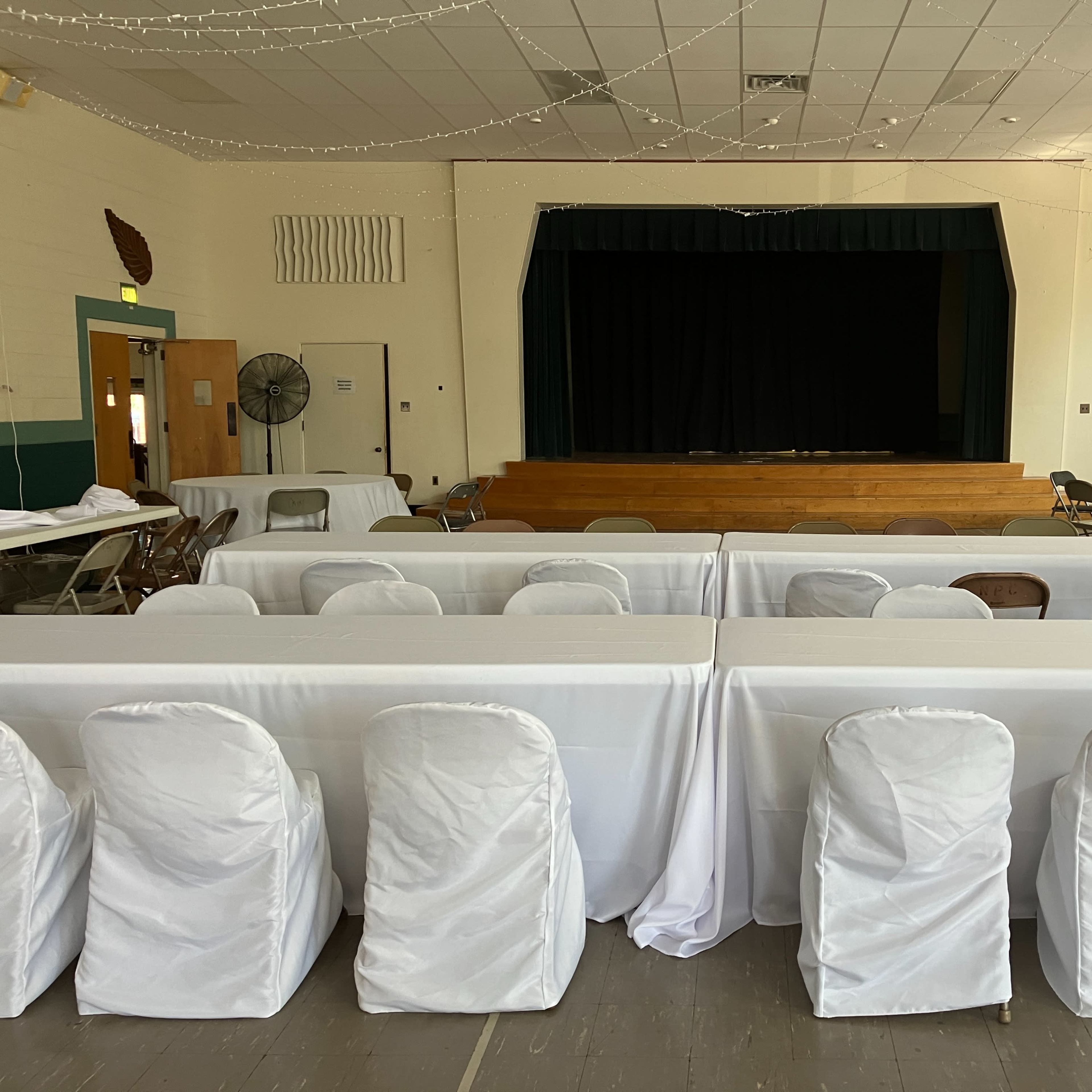 A spacious indoor event venue with rows of tables covered in white tablecloths and chairs wrapped in white coverings, facing a stage with a black curtain.