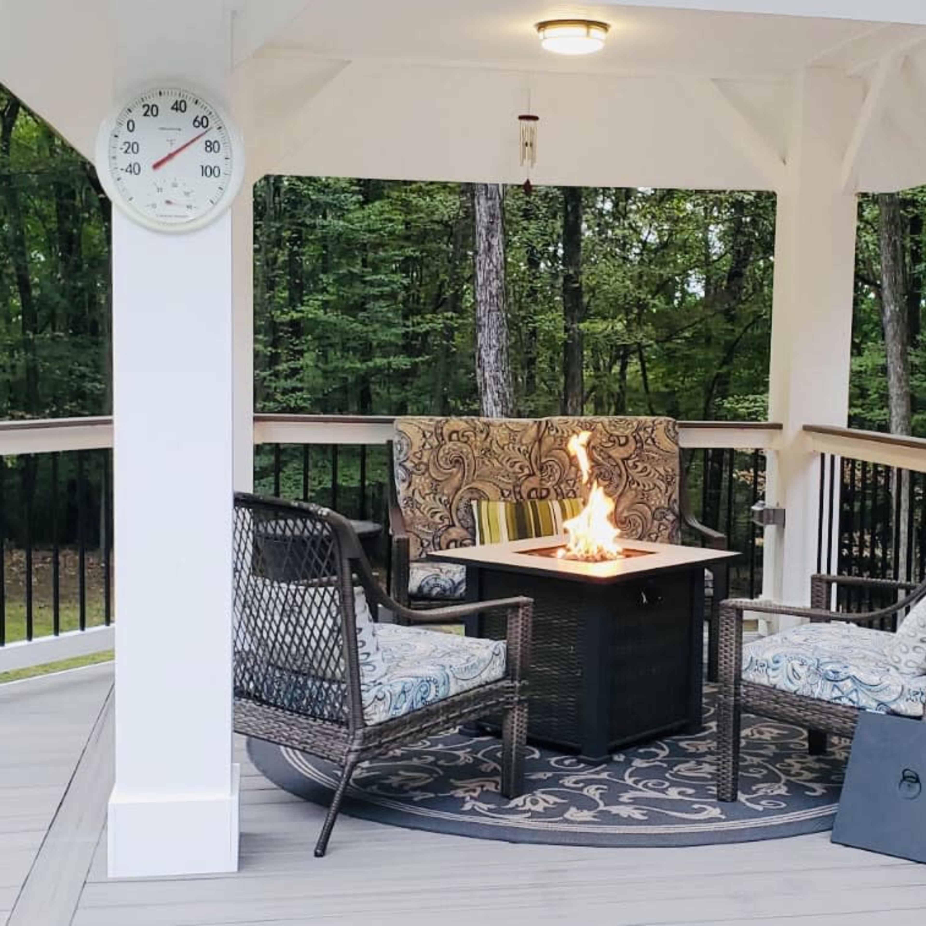 A gazebo features a table with a fire pit surrounded by two chairs on a deck overlooking a forested area.