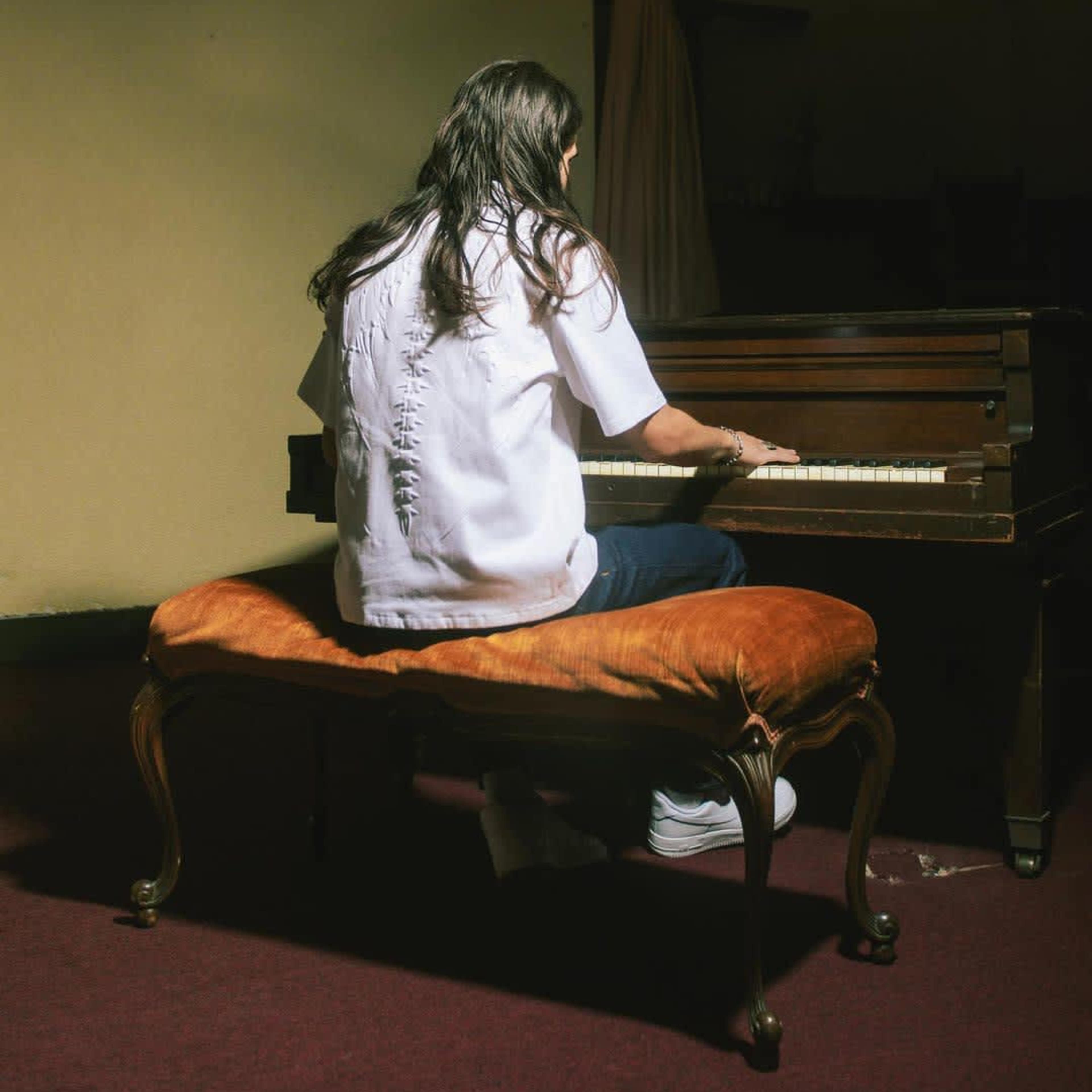 A person with long hair sits on a velvet cushion, playing a piano in a dimly lit room.