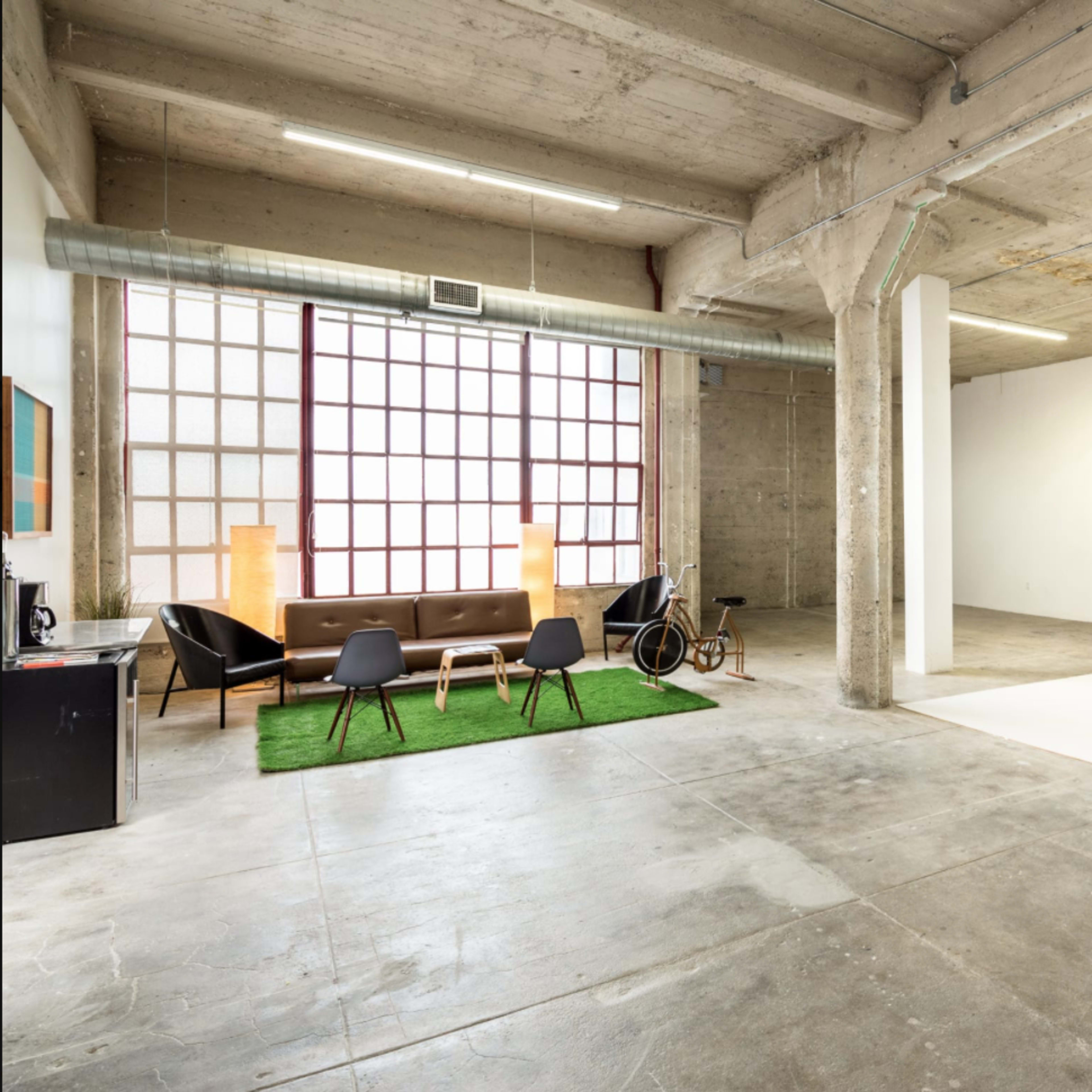 An industrial-style studio with concrete walls, a large window, a seating area with a sofa and chairs on a green rug, and an empty section featuring a desk with a computer.
