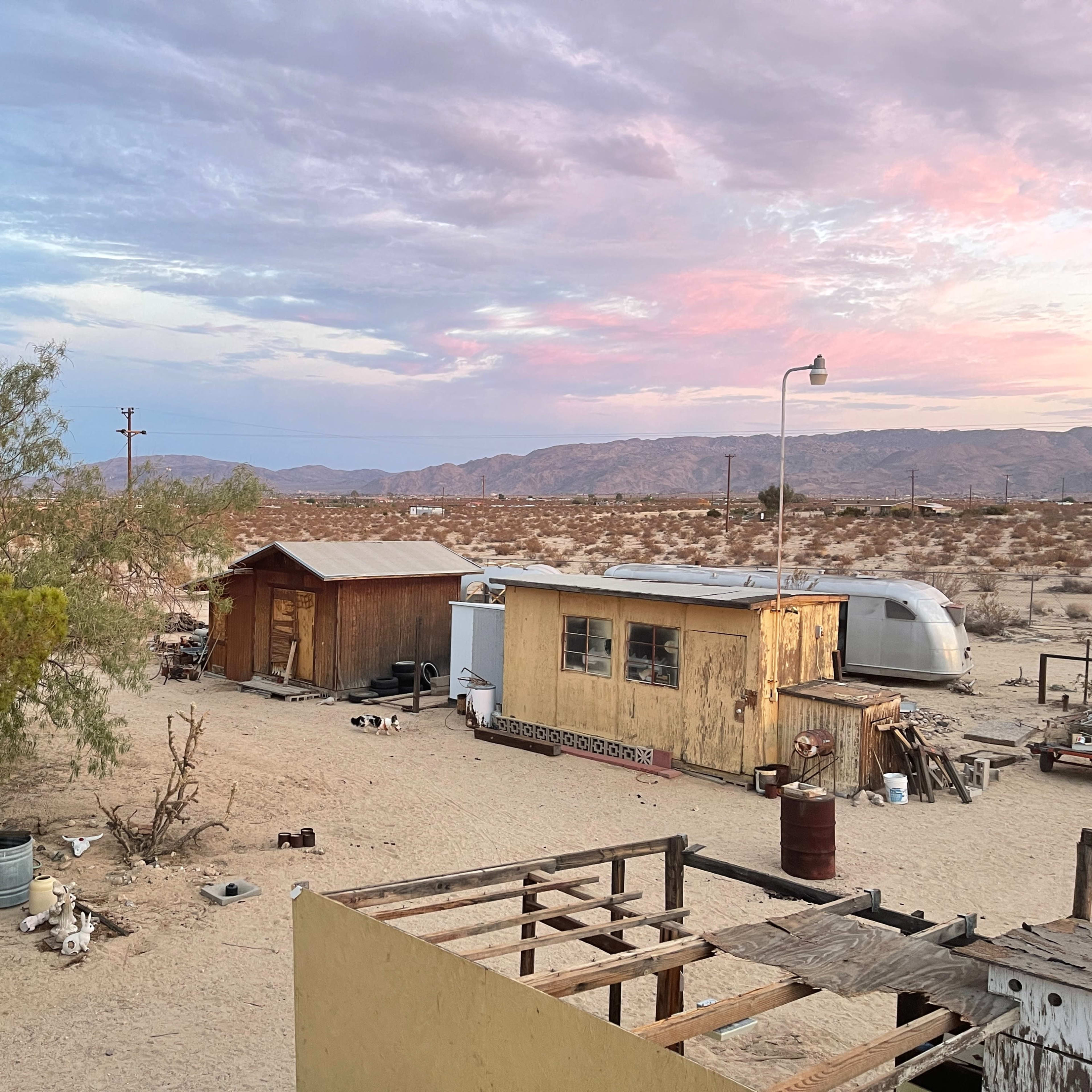 The scene shows a rustic desert landscape with a wooden shed, a trailer, and white outdoor chairs surrounded by dry ground and sparse vegetation.