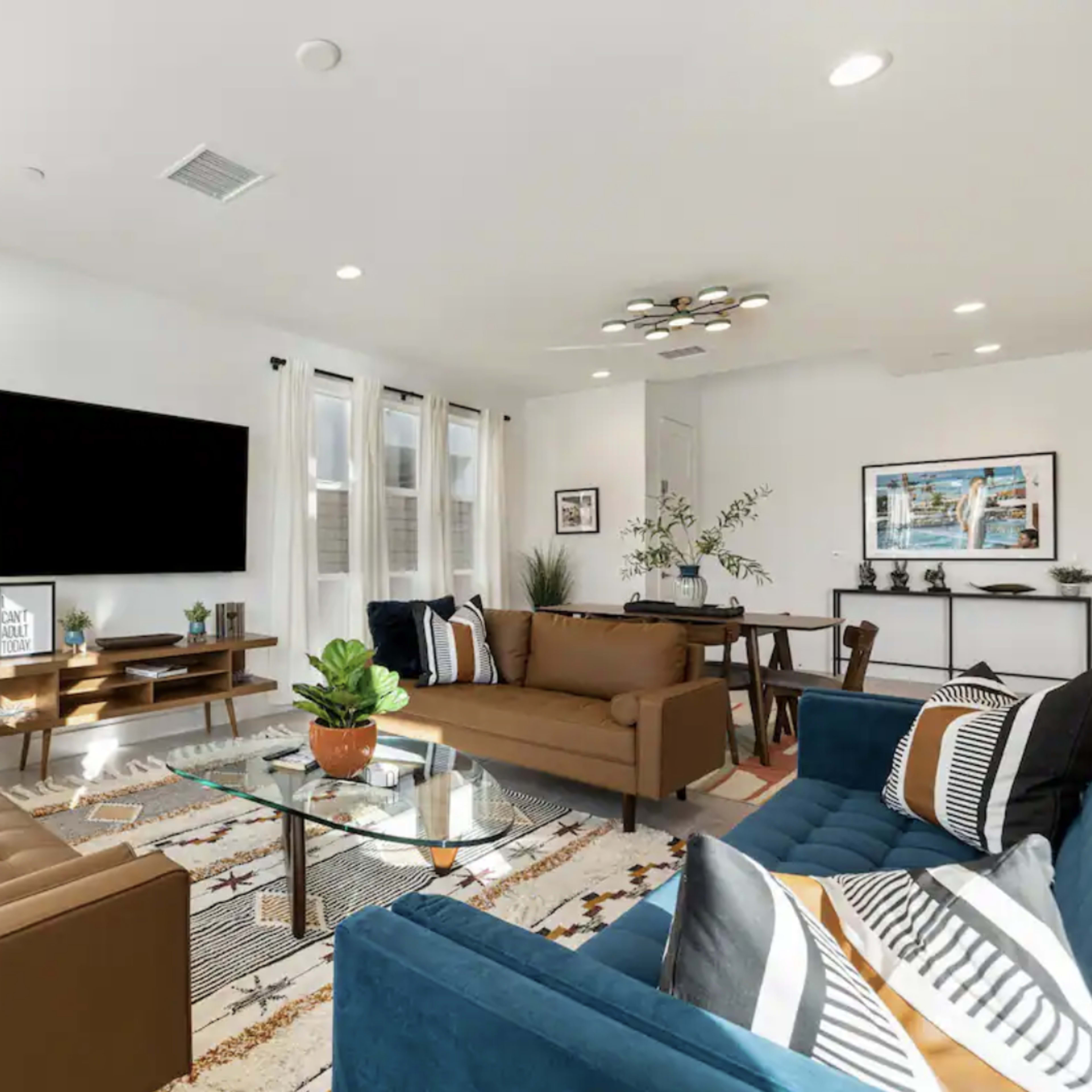 A modern living room with two sofas, a glass coffee table, a mounted television, and a dining area with a small table, all arranged around a patterned area rug.