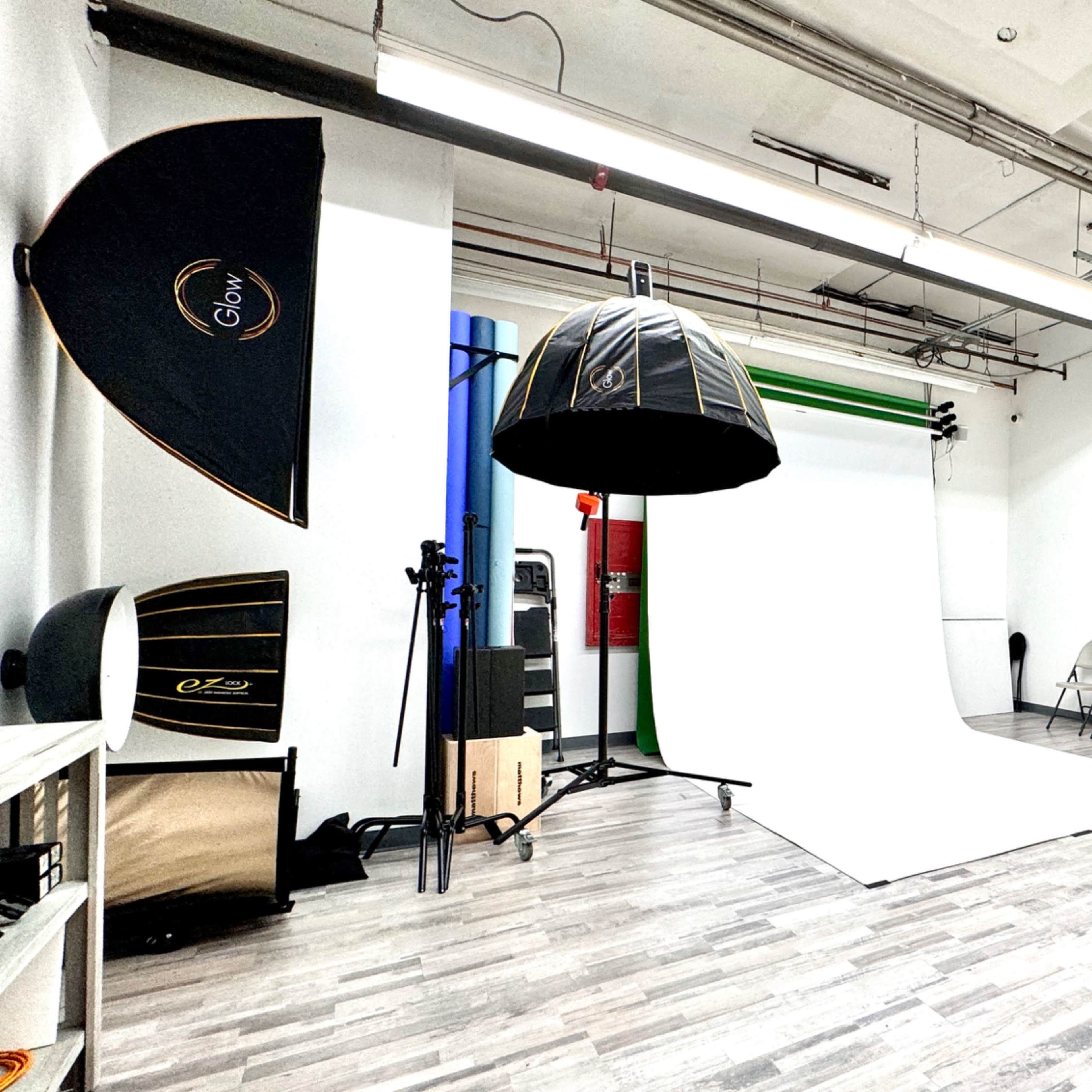 A photography studio with a white backdrop, lighting equipment, a director's chair, and various props arranged neatly in the space.