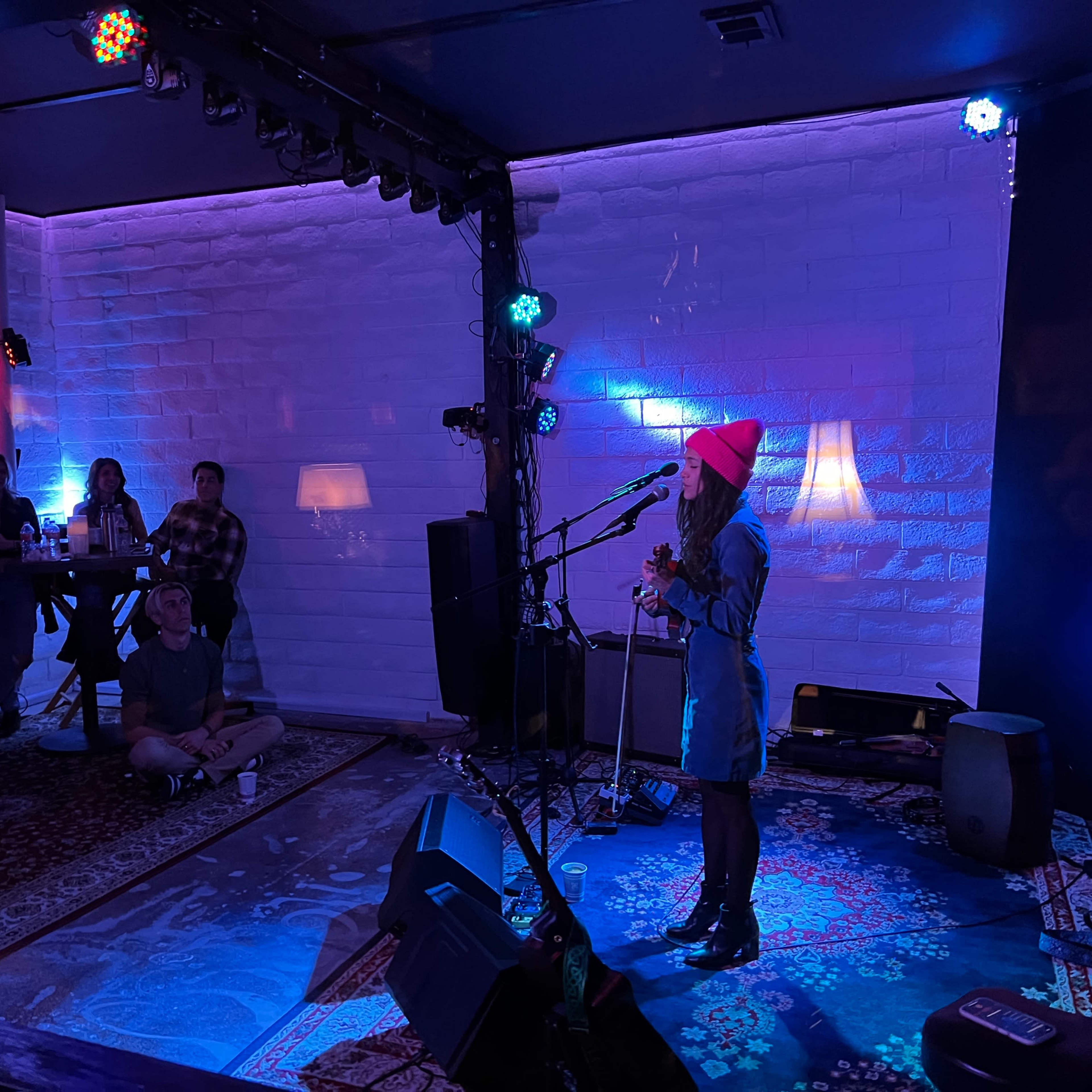 A performer stands on stage with a microphone in front of an audience seated in a dimly lit venue adorned with colorful lights.