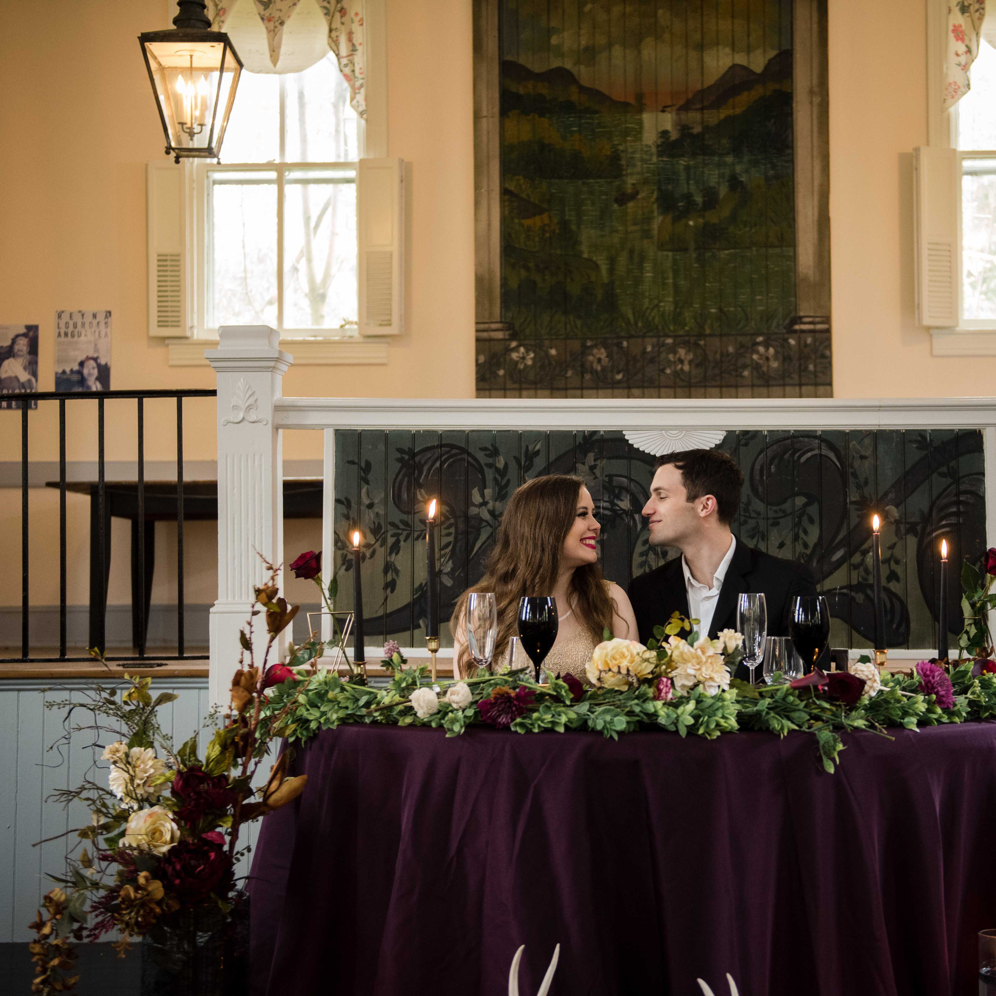 A couple sits at an elegantly decorated table with flowers and candles in a warmly lit indoor space.