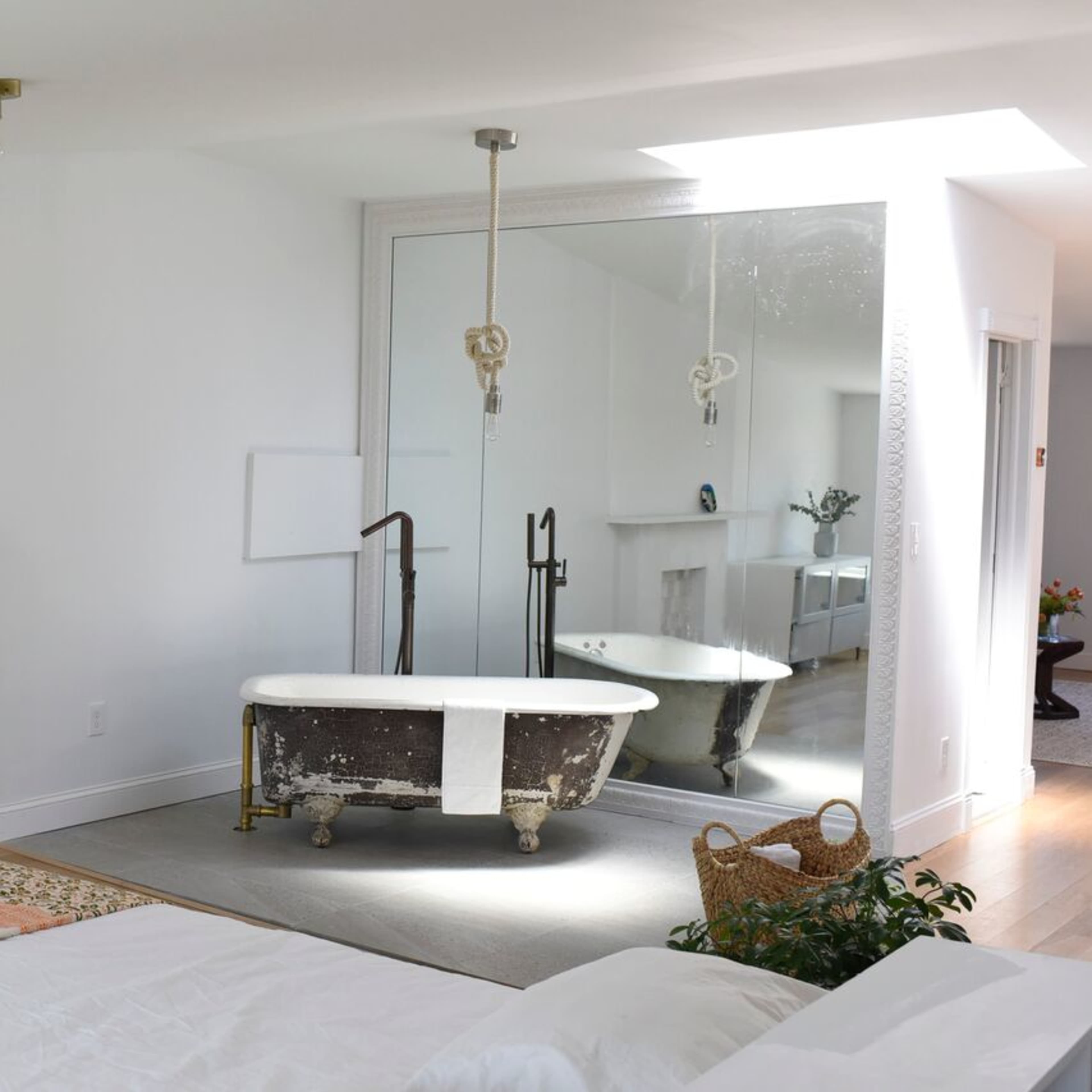 A minimalist bedroom featuring a freestanding bathtub reflected in a large mirror, with a neutral color palette and a decorative rug on the floor.