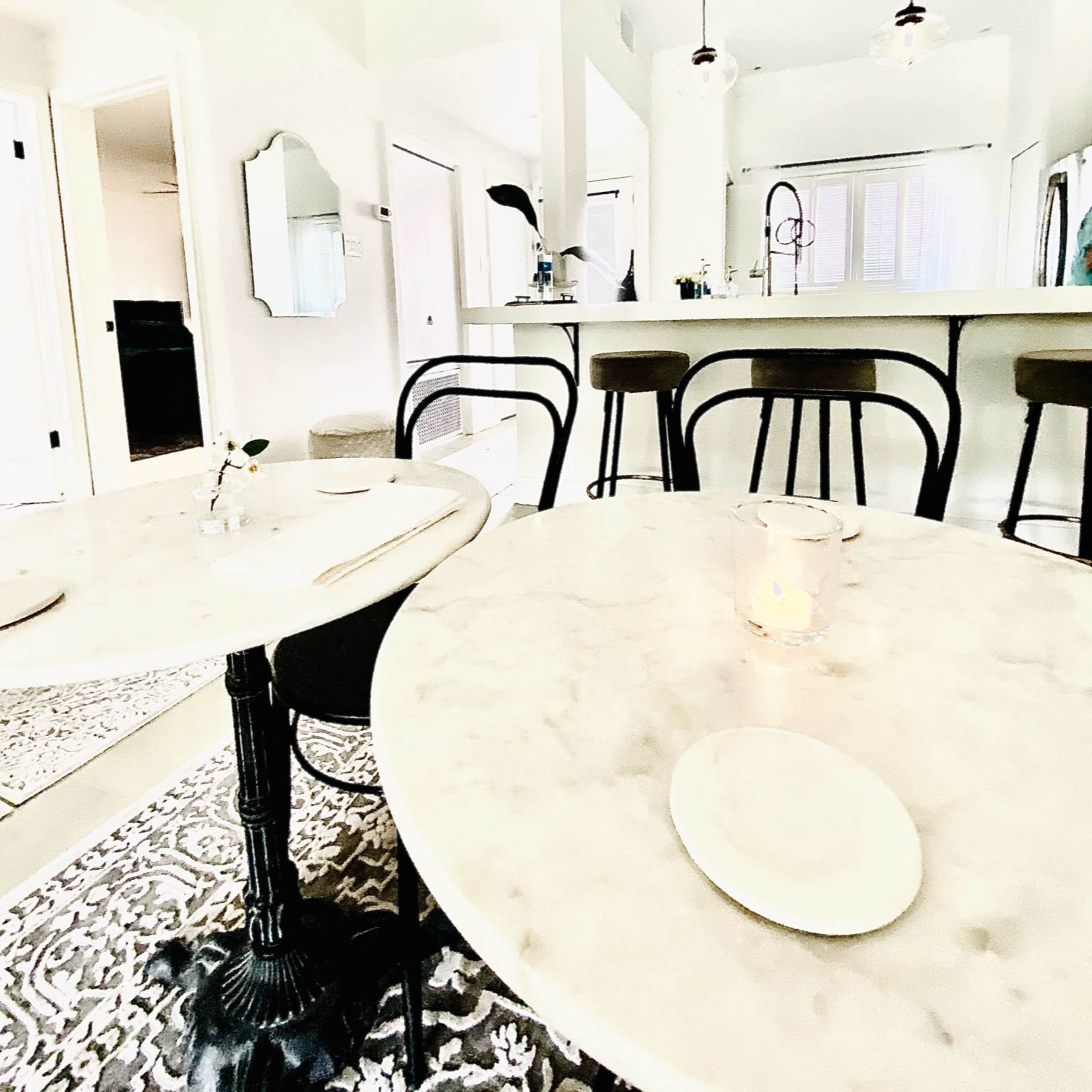 A bright restaurant interior featuring marble-topped tables, black chairs, and a kitchen visible in the background.