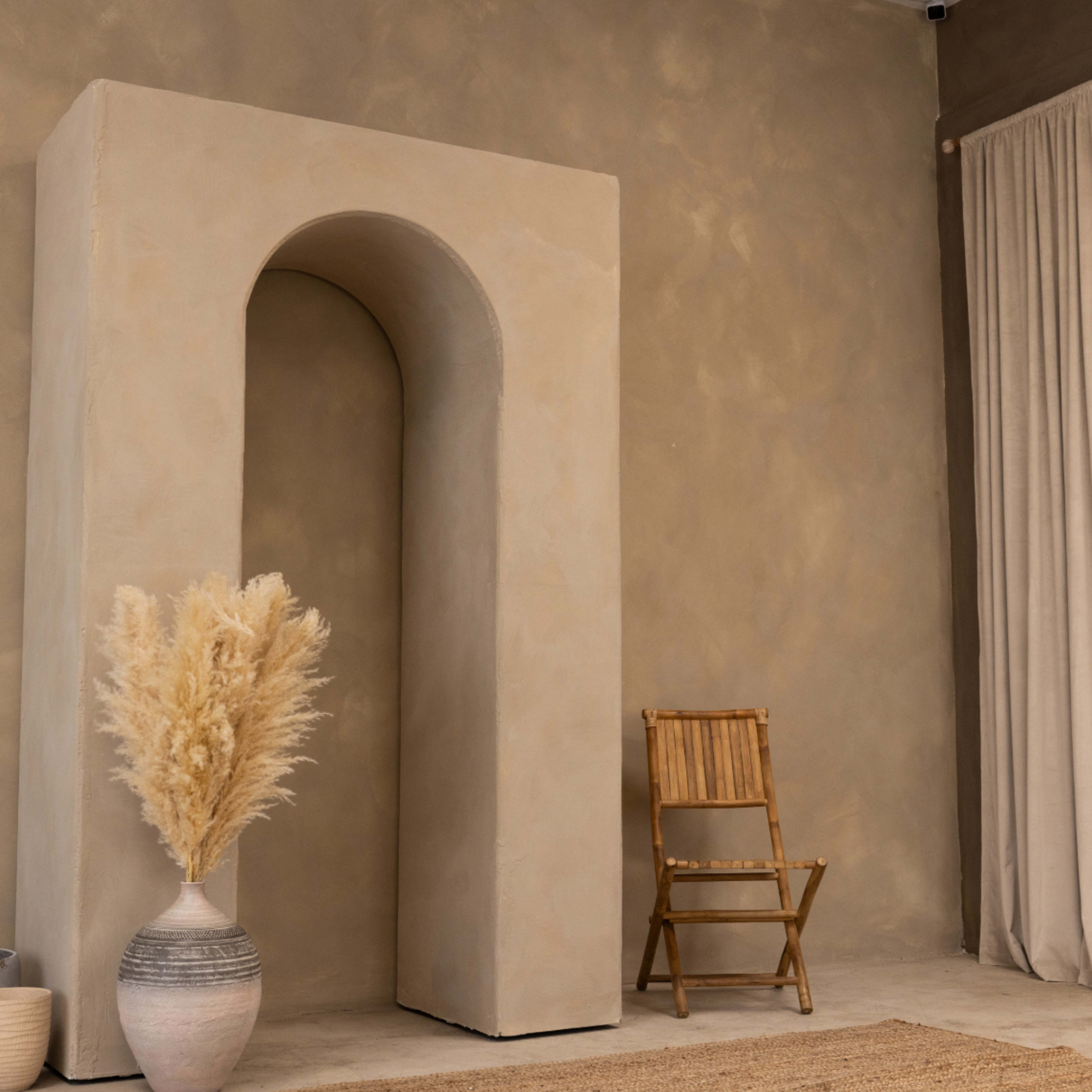 A minimalist interior space featuring a large archway, a vase with dried grasses, a wooden chair, and neutral-colored walls and curtains.