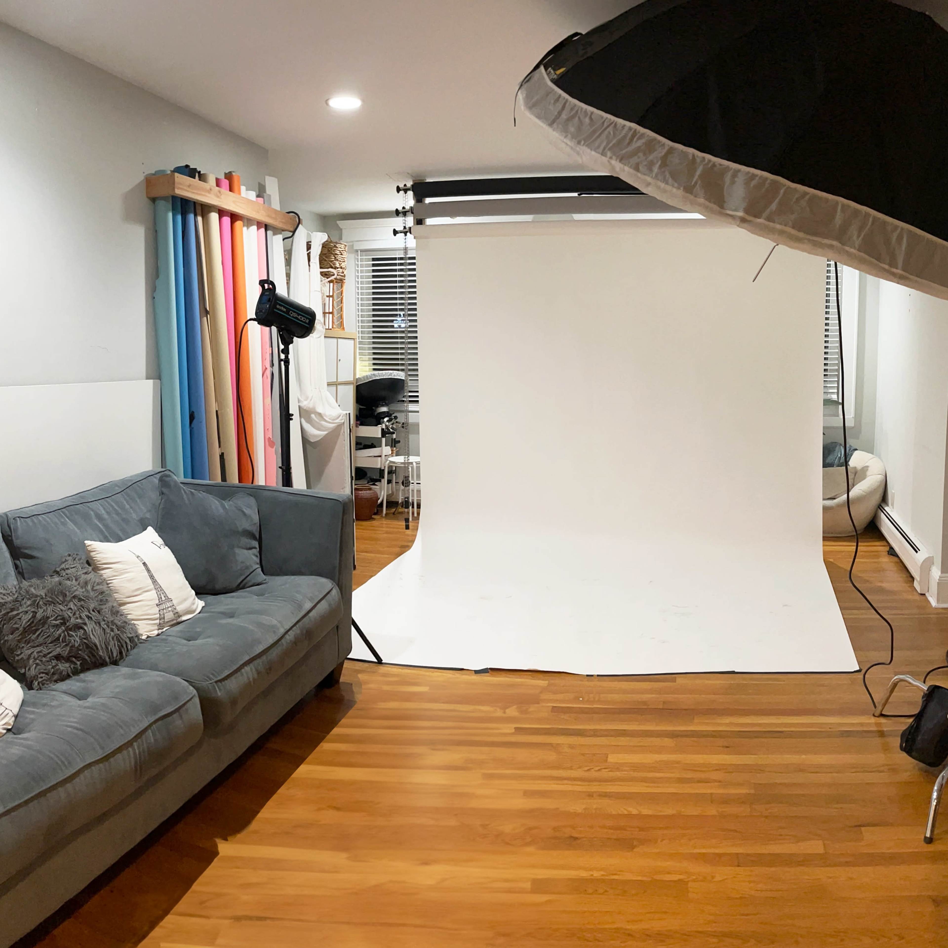 A photography studio features a gray couch, a plain white backdrop, and various lighting equipment.
