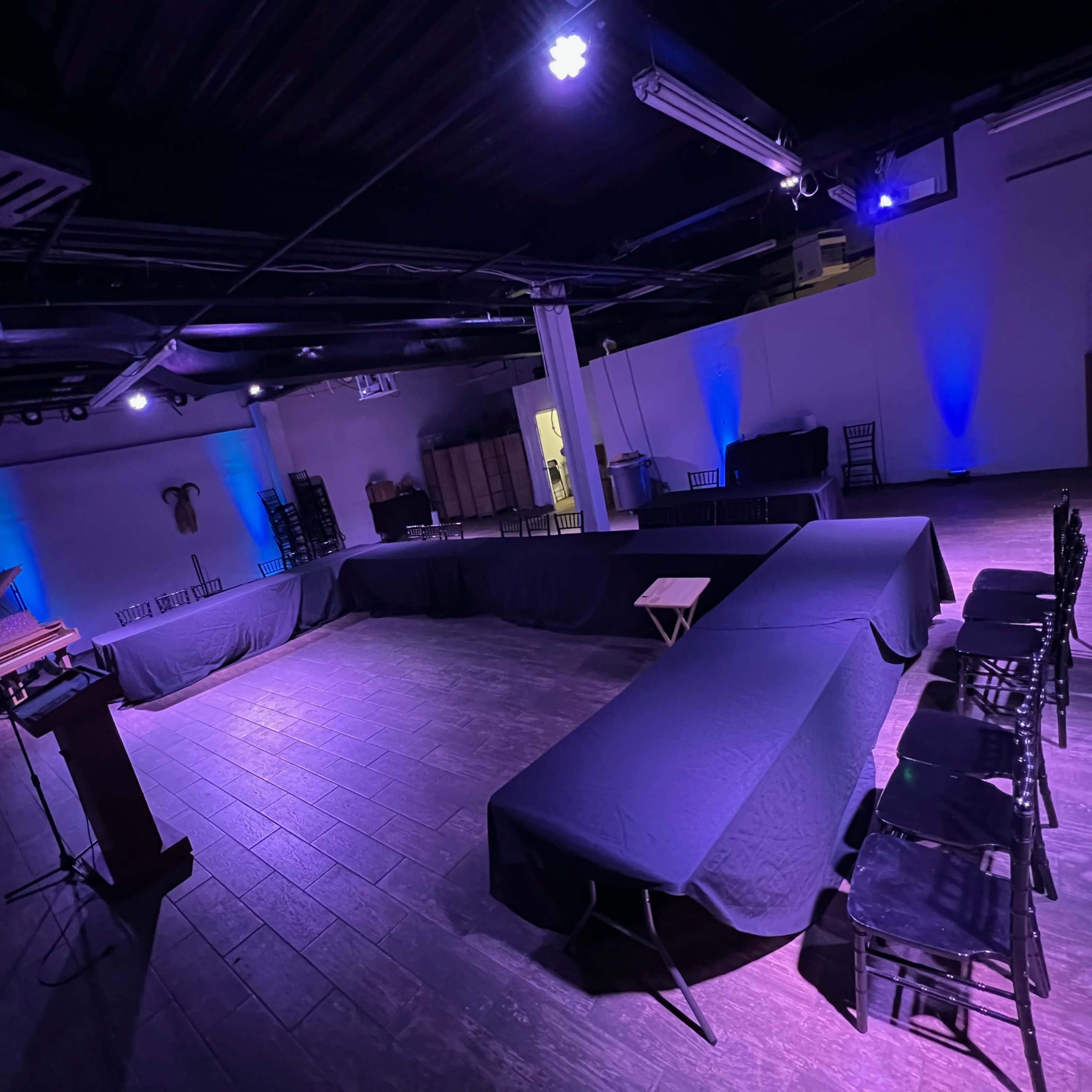 A spacious event venue with tables covered in black cloth, arranged in a U-shape, and dimly lit with blue and purple lighting.