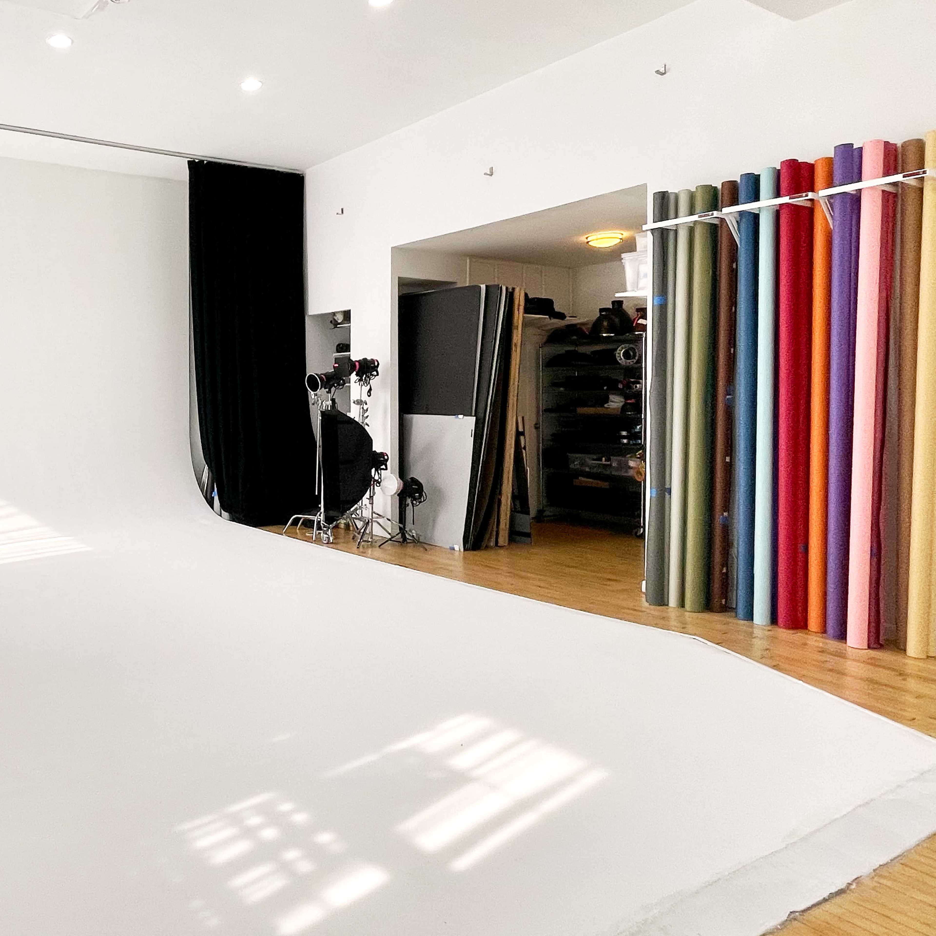 A spacious photography studio with a white seamless backdrop, studio lights on the left, and a storage area featuring rolls of colored backdrops on the right.