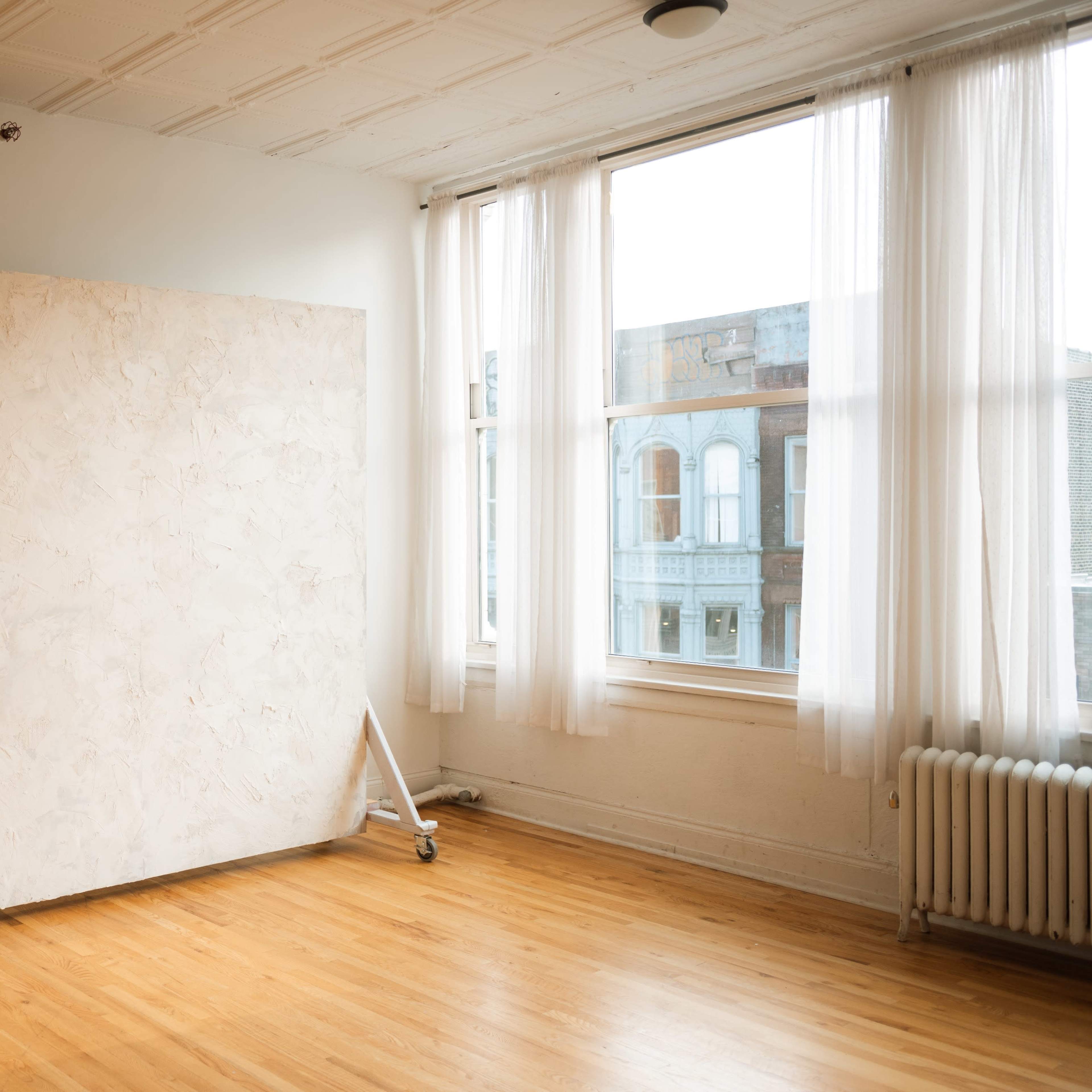 A spacious, well-lit room featuring a large, unpainted canvas on wheels positioned against a wall with large windows.