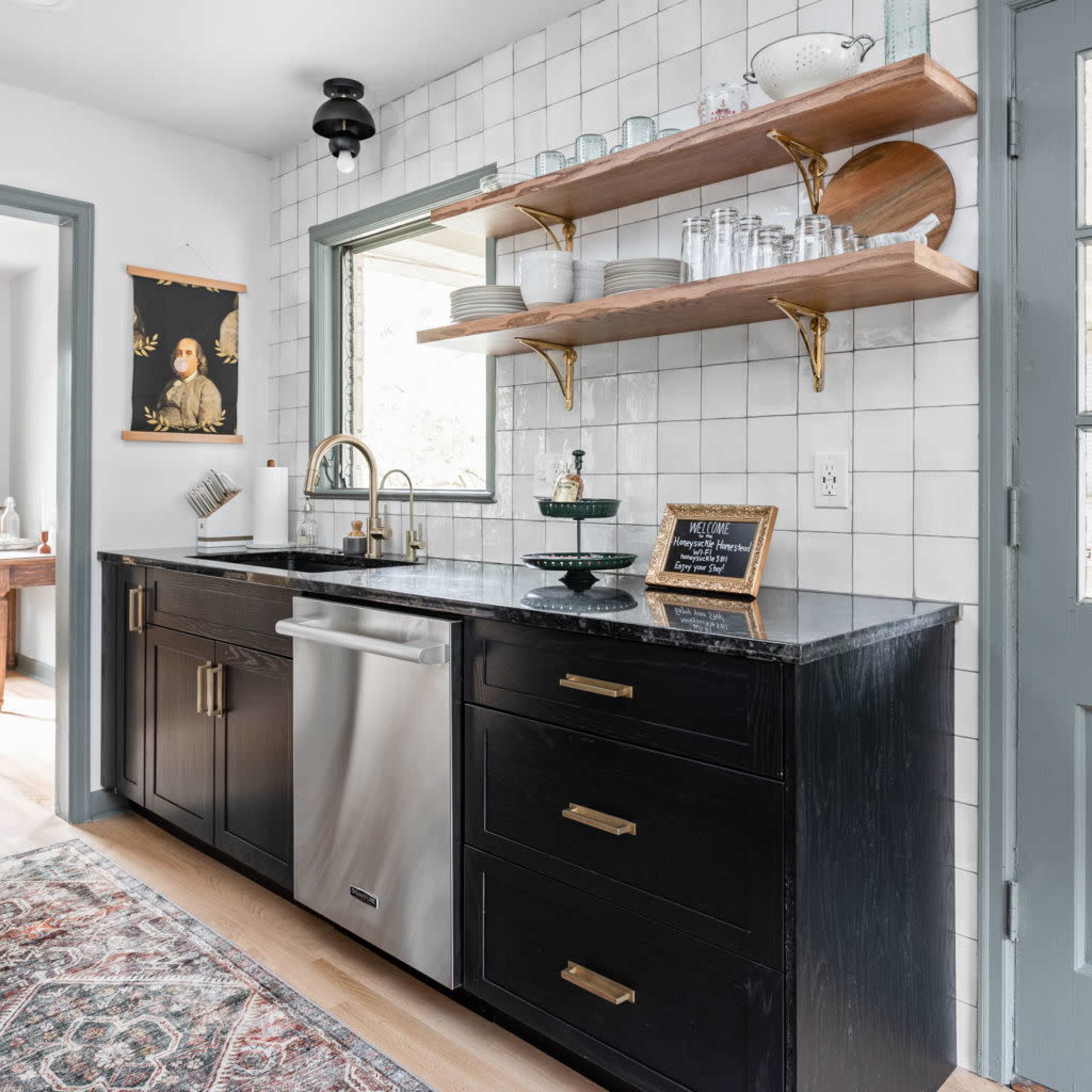 A modern kitchen featuring black cabinetry, a stainless steel sink, and open wooden shelving with glassware against a tiled wall.