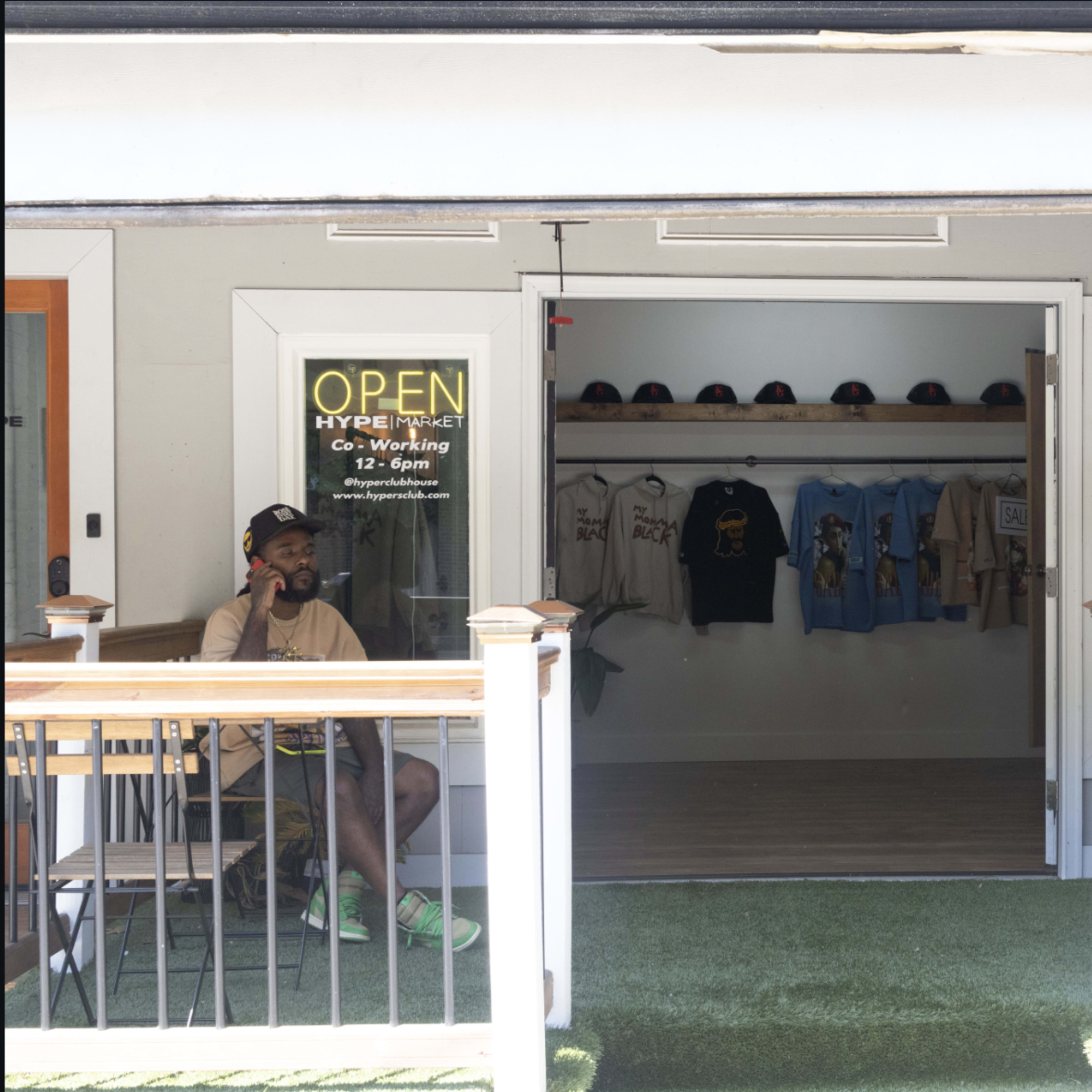 Two men sit at a table outside a storefront labeled "OPEN" and "HYPE MARKET," which displays clothing and artwork inside.