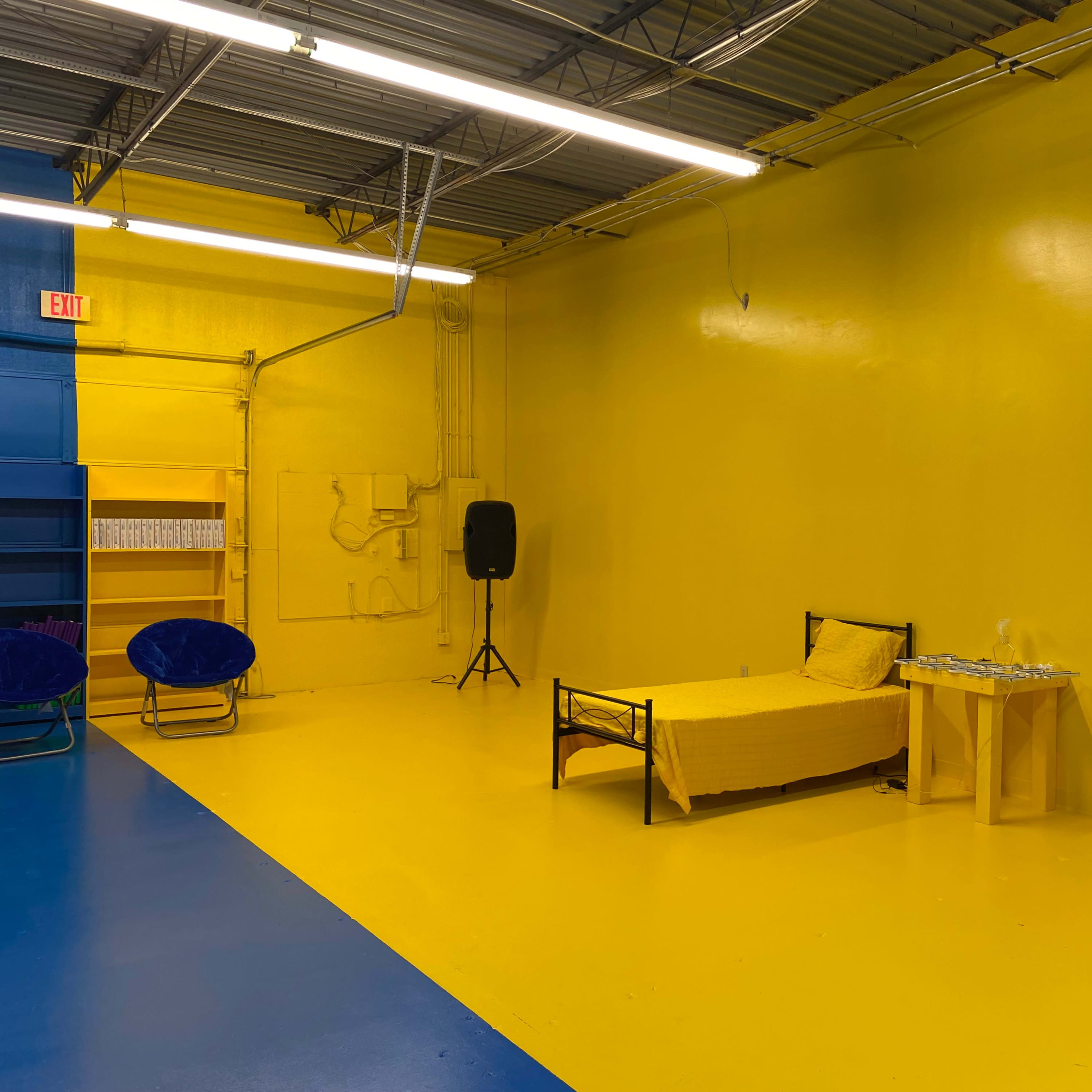 A room with yellow walls and flooring, featuring a bed with a yellow bedspread, a small table, and a blue area on one side.