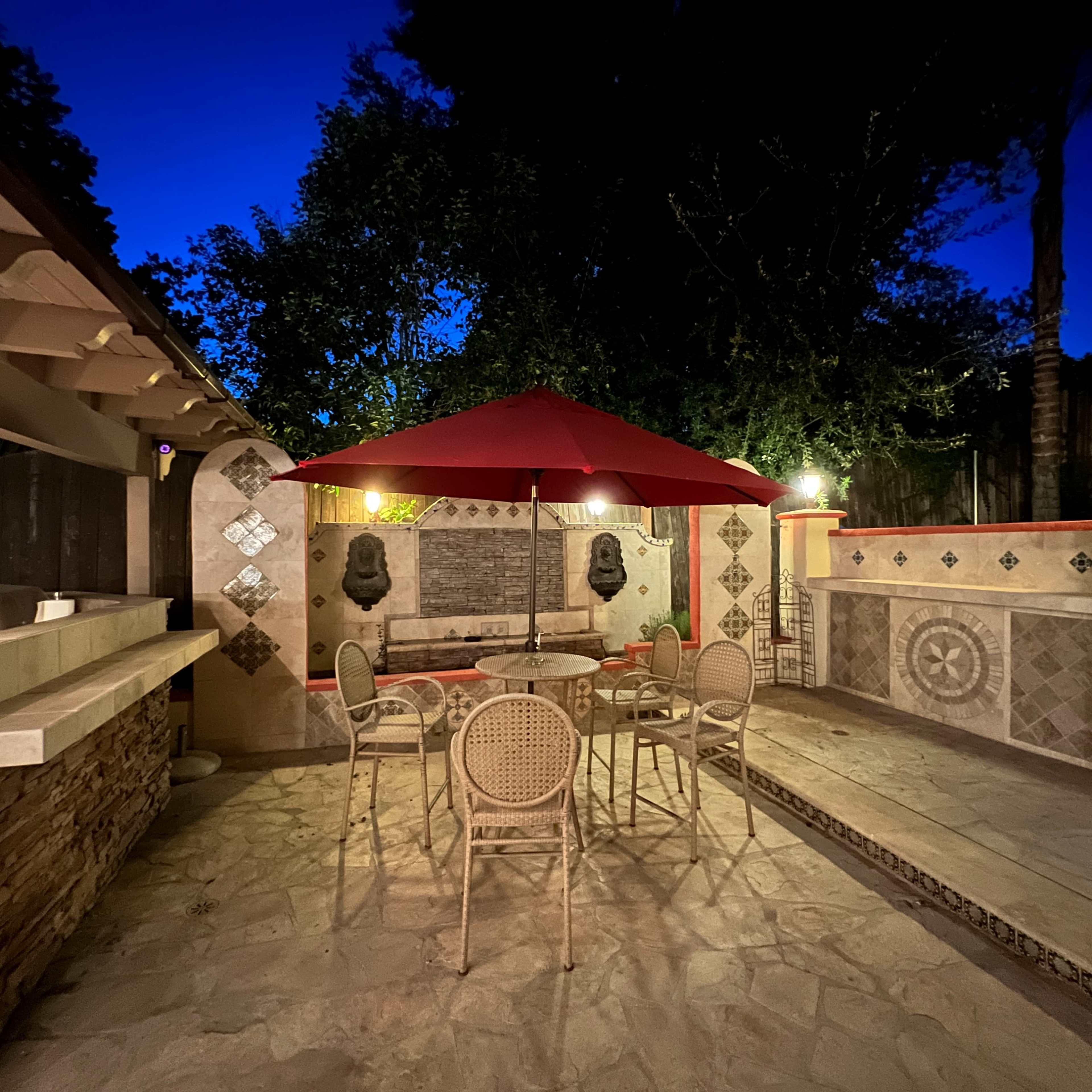 A patio area features a table and chairs under a red umbrella, with a stone wall and outdoor kitchen in the background.