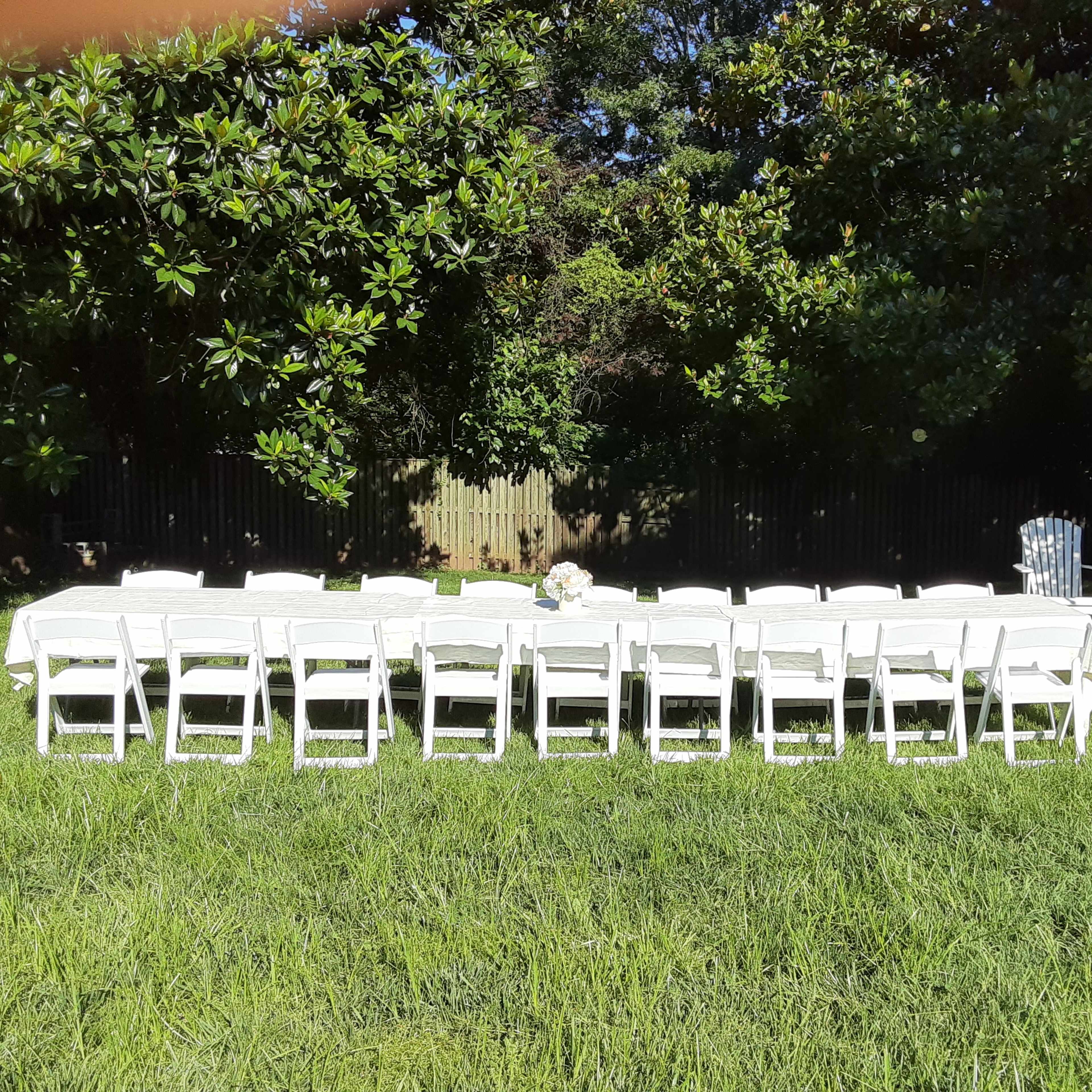 A long table with white tablecloths and chairs is set up on a grassy area surrounded by trees.