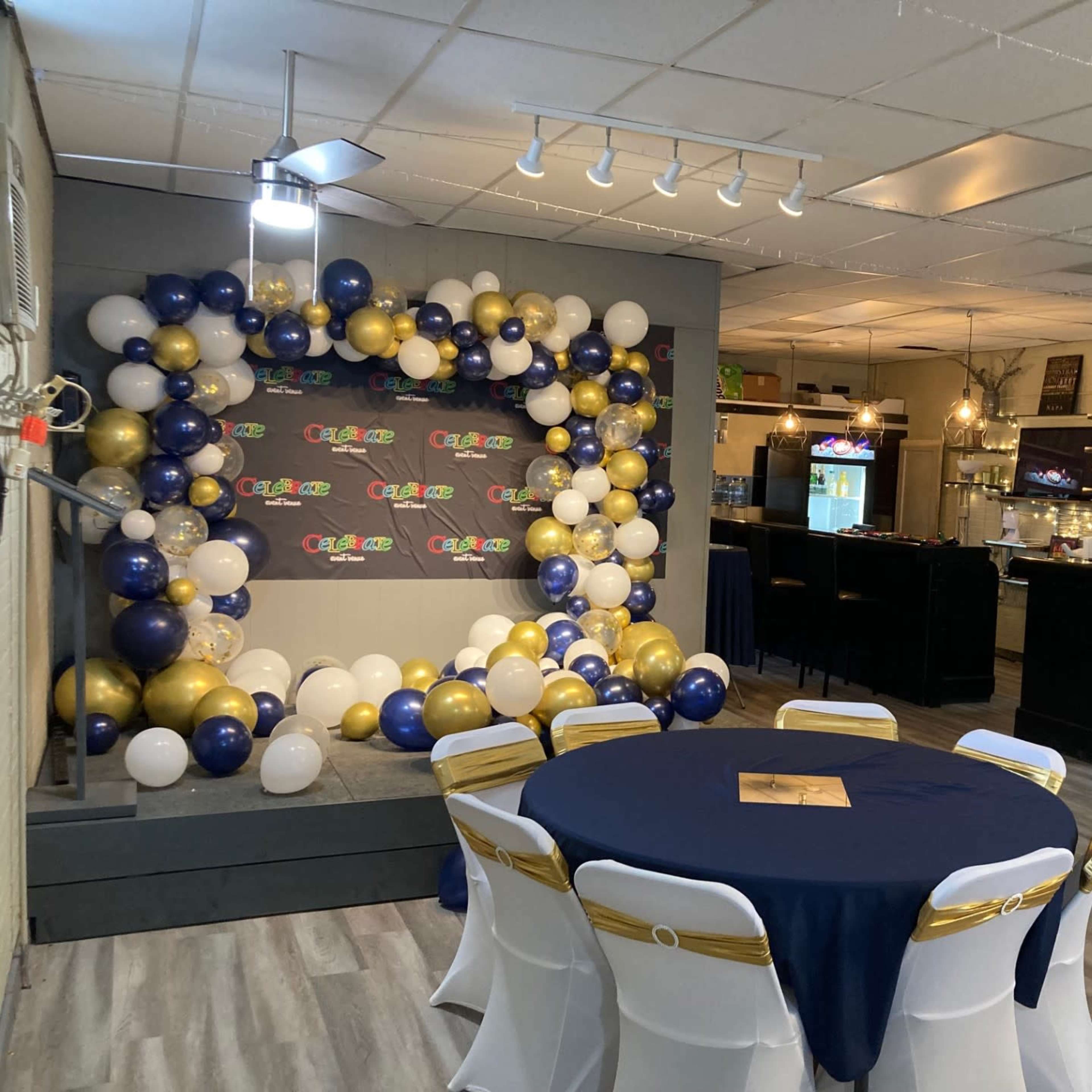 A decorated event space featuring a balloon arch, tables with dark blue tablecloths, and a bar area in the background.