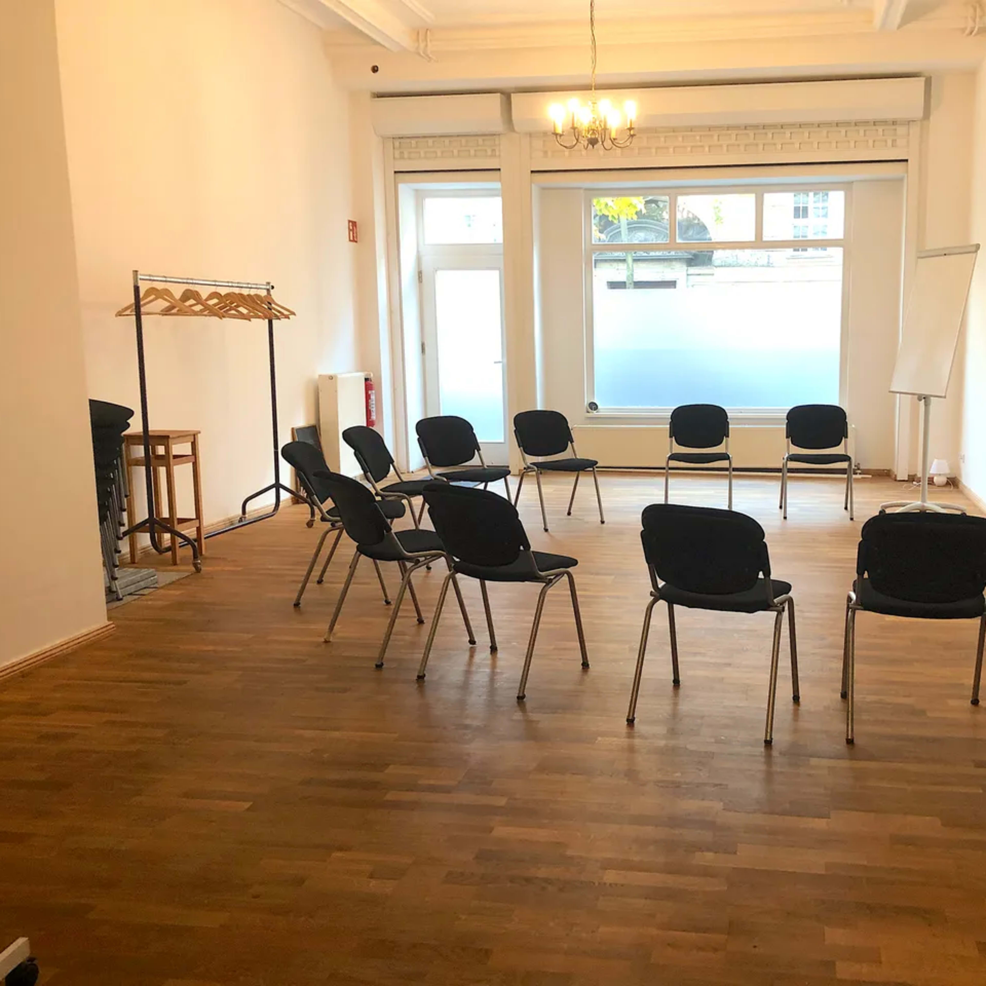 A bright room with wooden flooring features a circle of chairs, a whiteboard, and a coat rack.
