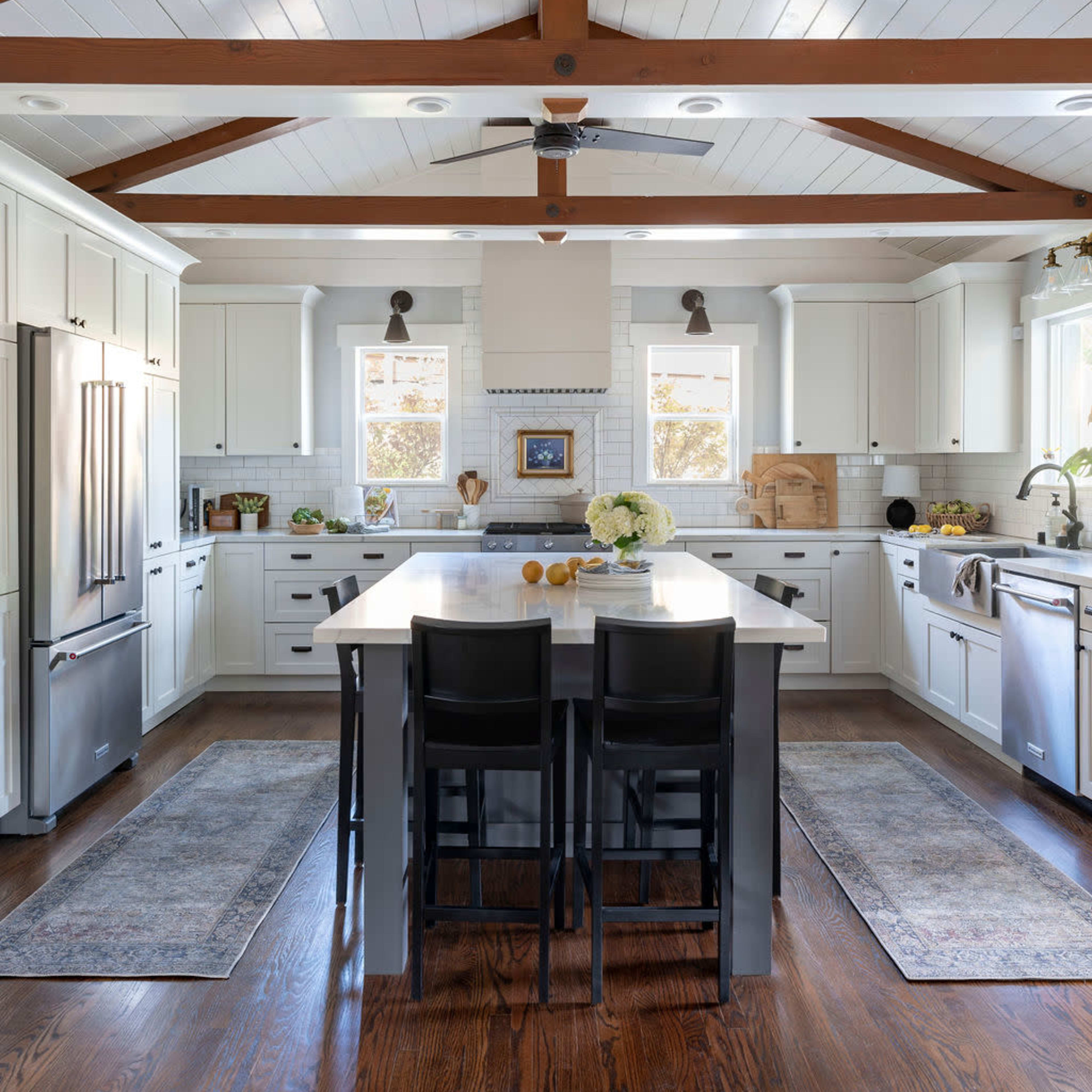 A modern kitchen features white cabinetry, a large island with dark seating, and wooden ceiling beams.