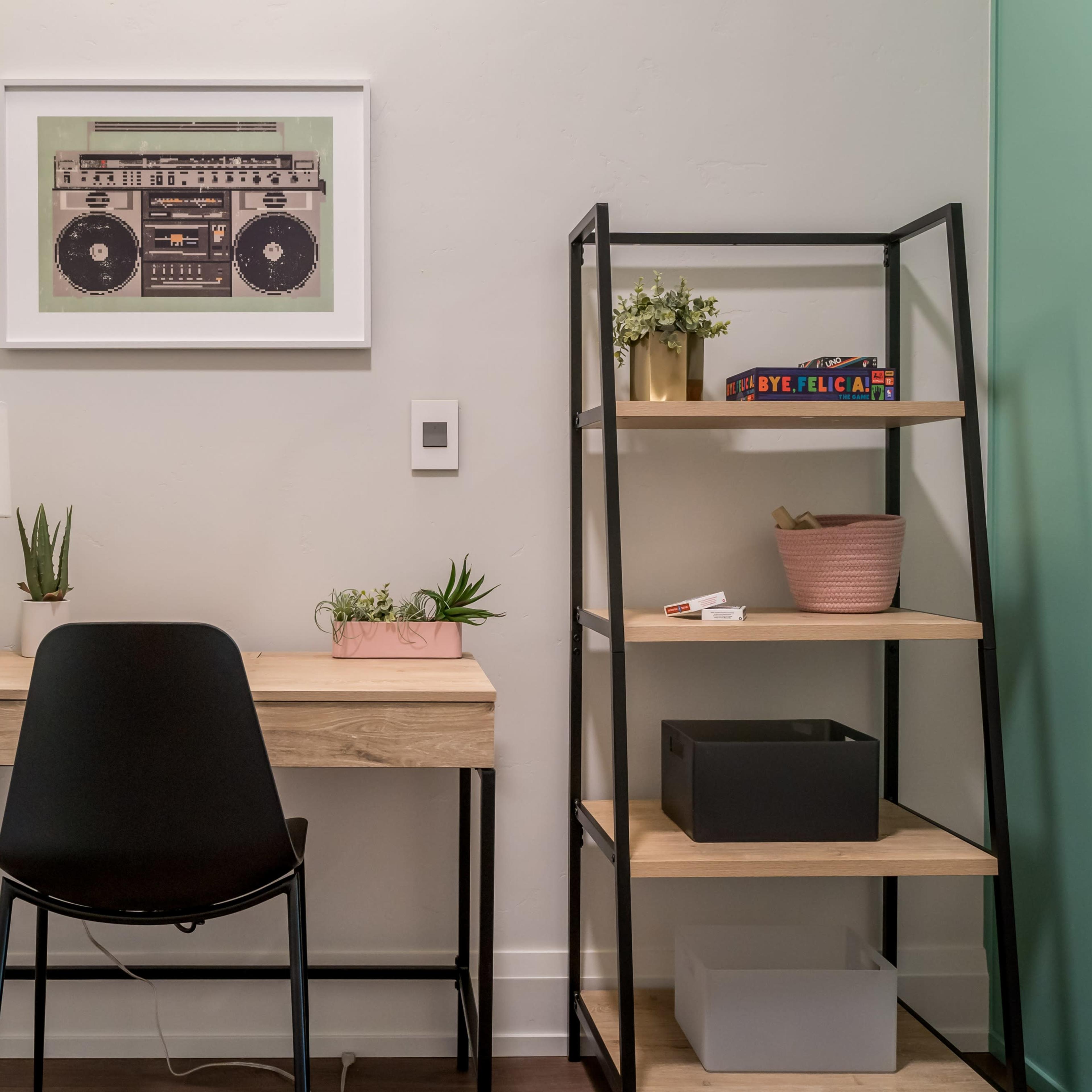 A minimalist workspace featuring a wooden desk with a chair, a lamp, and decorative plants, alongside a black shelving unit containing various items.