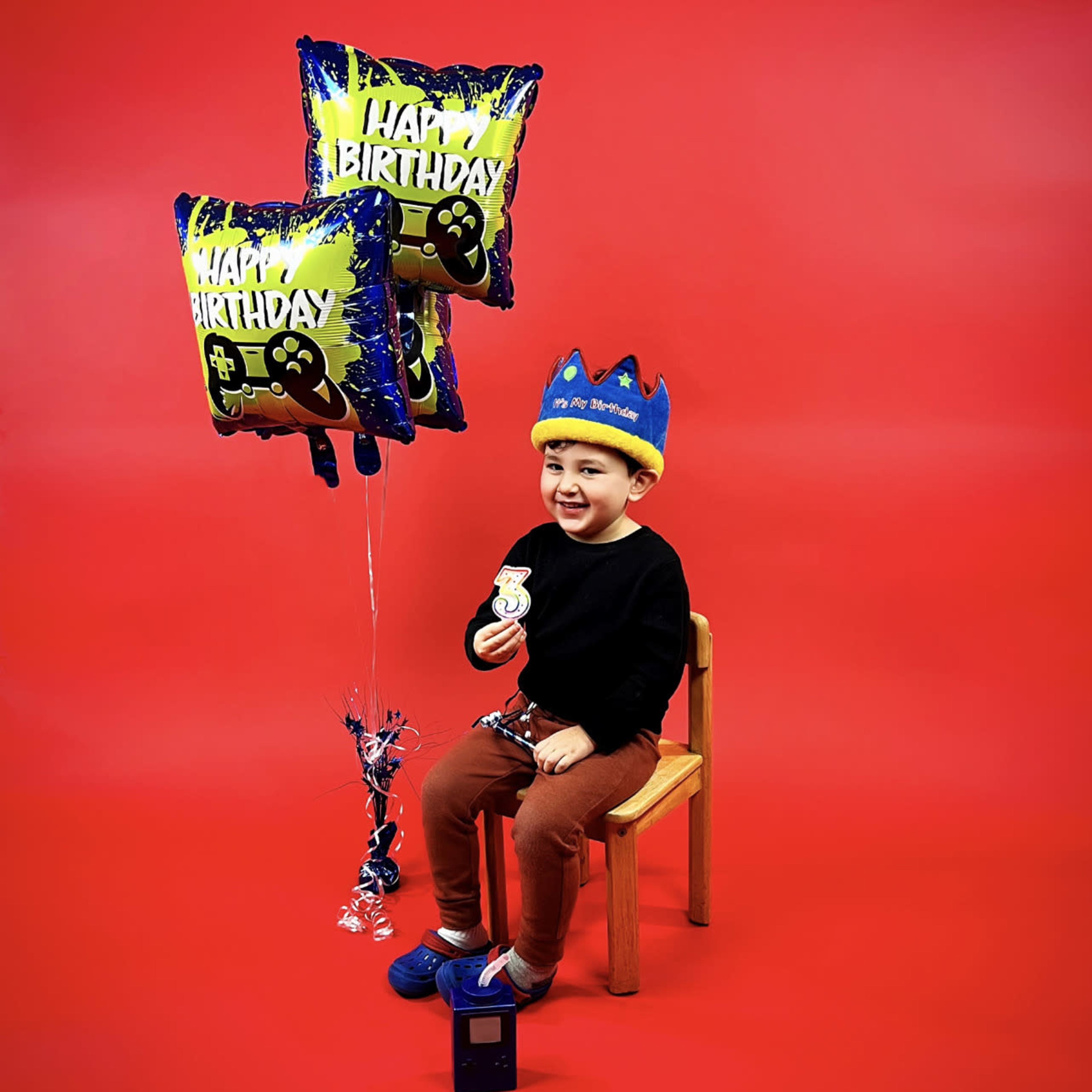 A young child sits on a wooden chair holding birthday balloons and a cupcake against a red background.