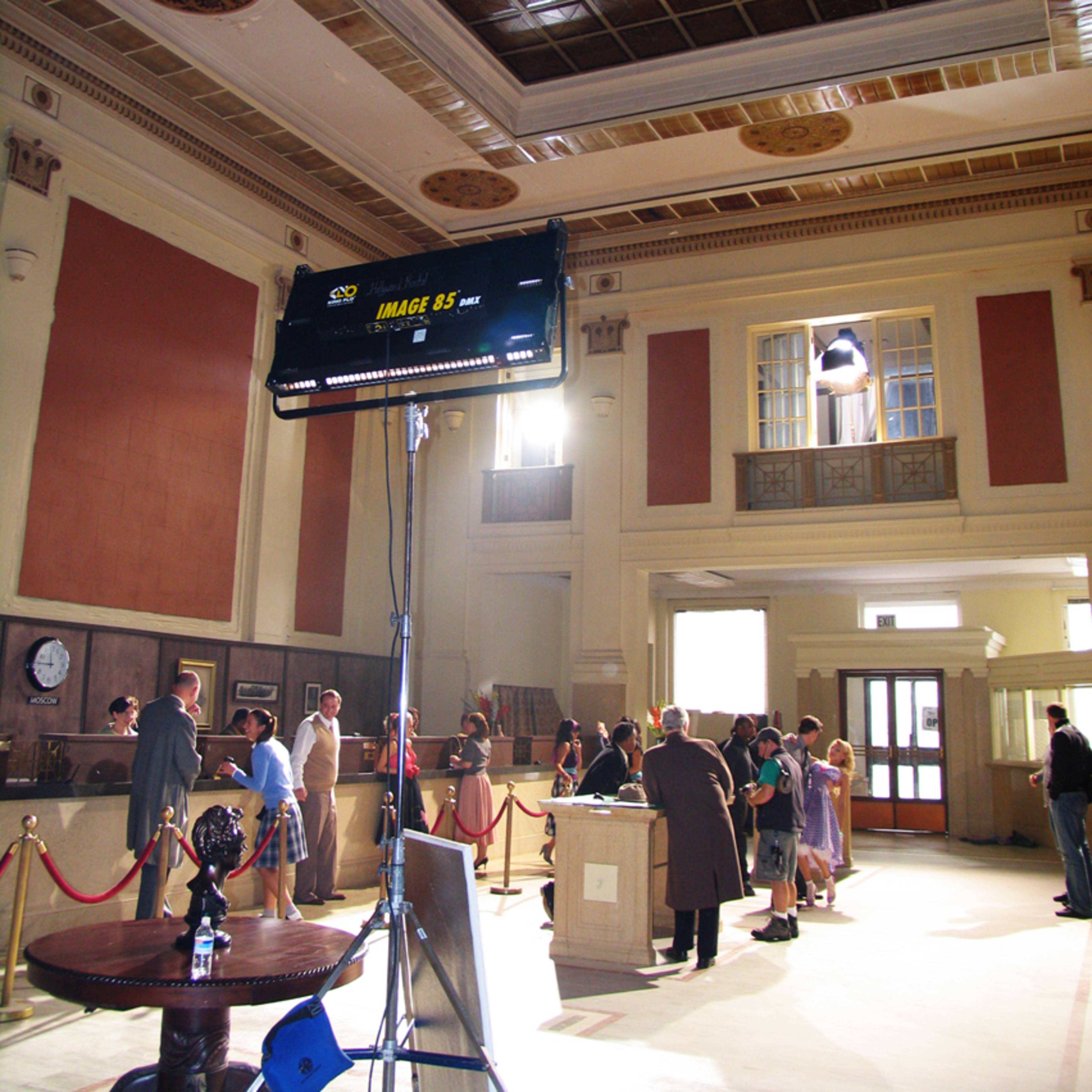 A film crew sets up a scene inside a grand, partially lit hall featuring high ceilings and decorative architecture.