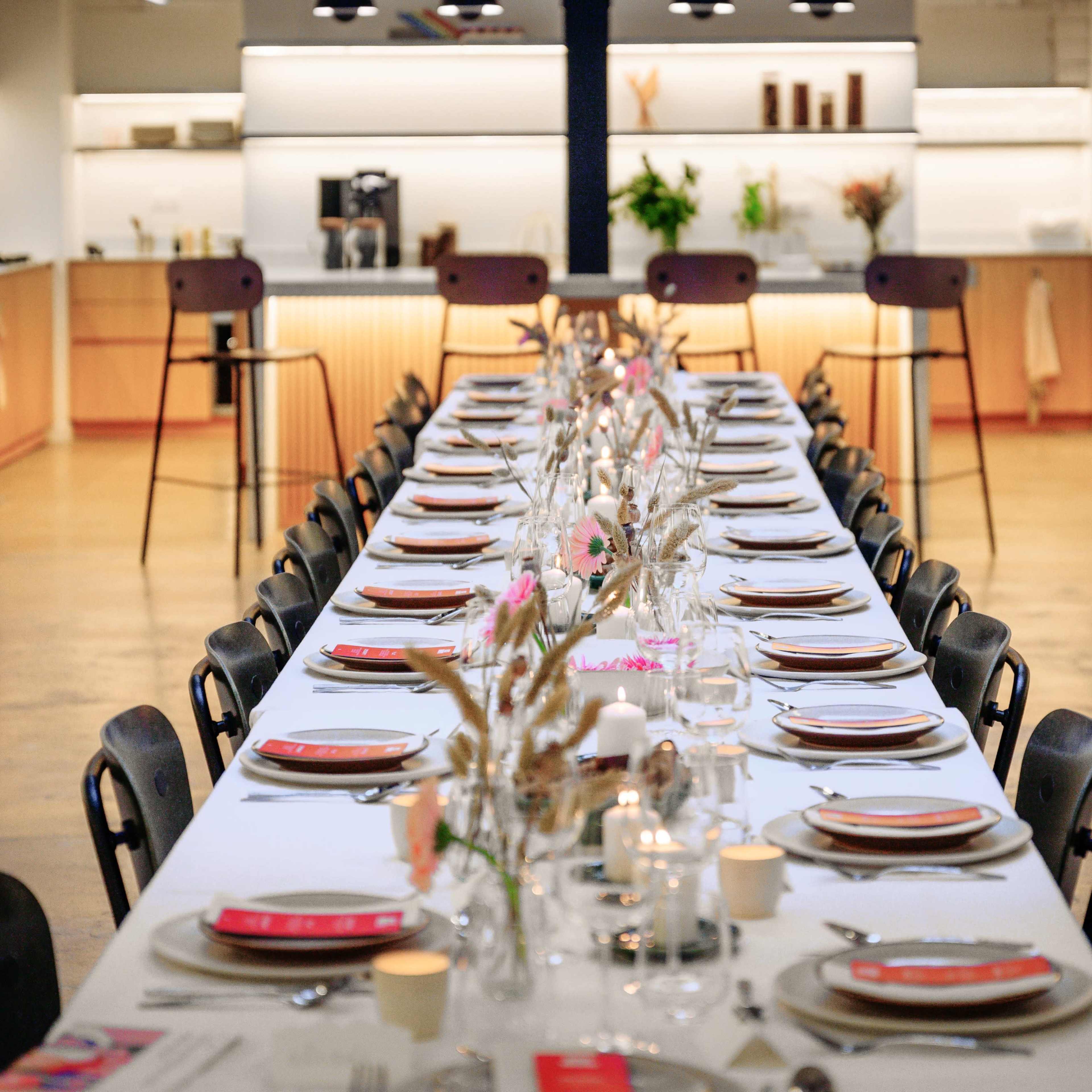 A long dining table is set with plates, glasses, and decorative centerpieces, surrounded by chairs in a bright, modern kitchen space.