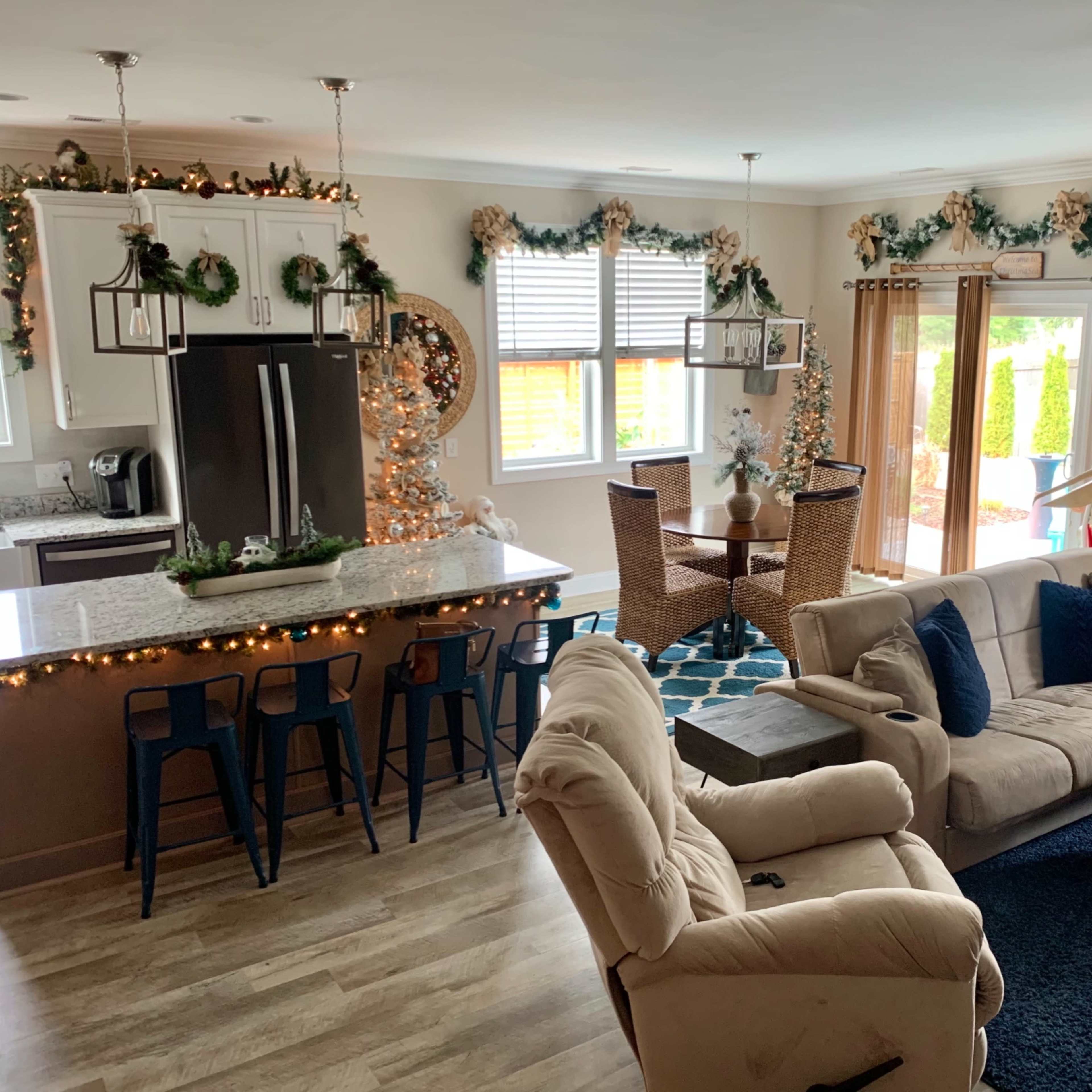 A well-decorated open-concept living space features a kitchen with bar seating, a dining area with wicker chairs, and festive holiday decorations throughout.
