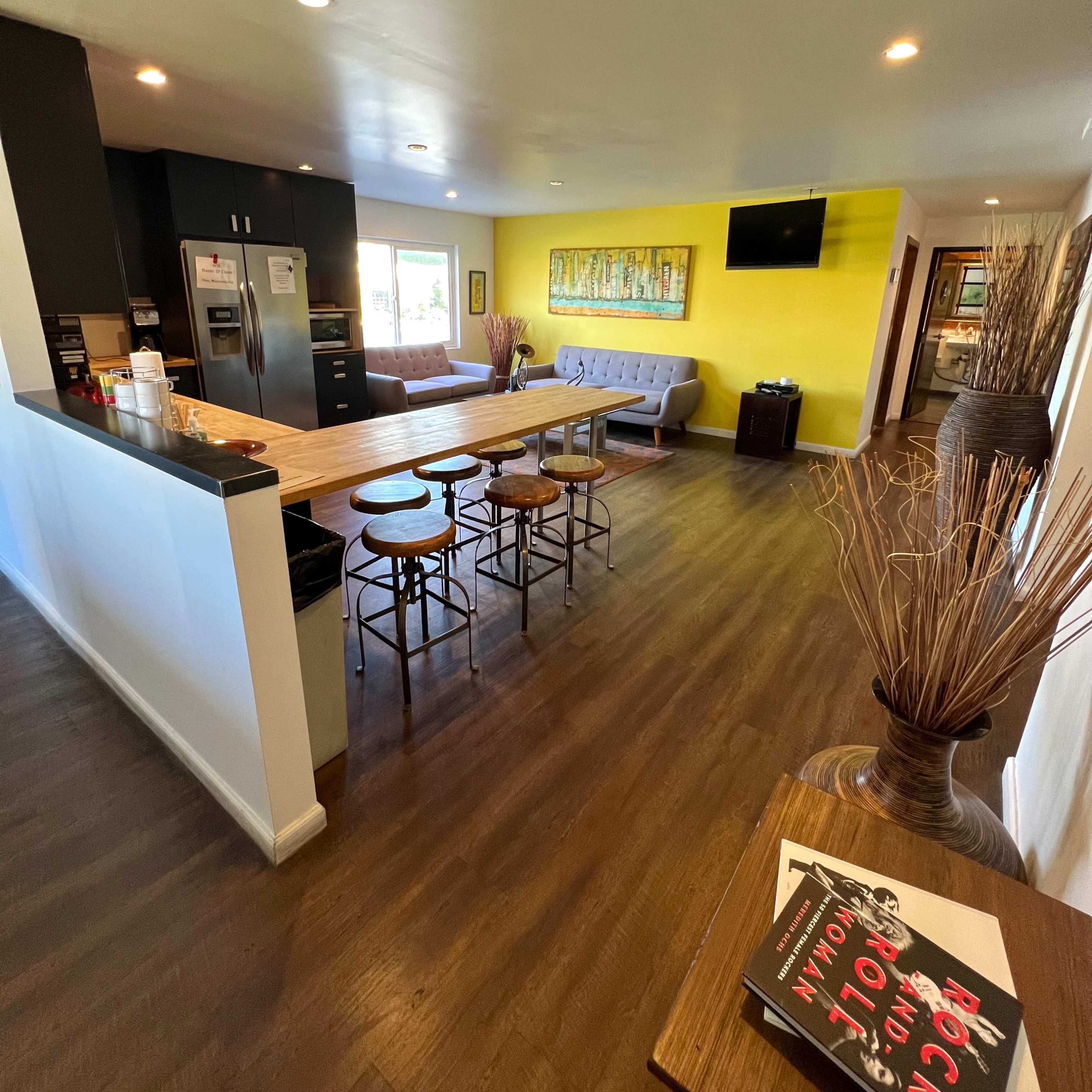 A modern open kitchen and dining area with a long wooden table, bar stools, and a bright yellow accent wall.